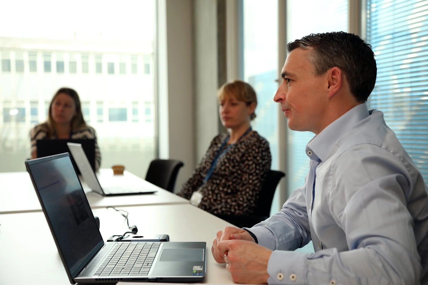 Businesspeople in a meeting room with laptops, sitting at a table, with large windows in the background.