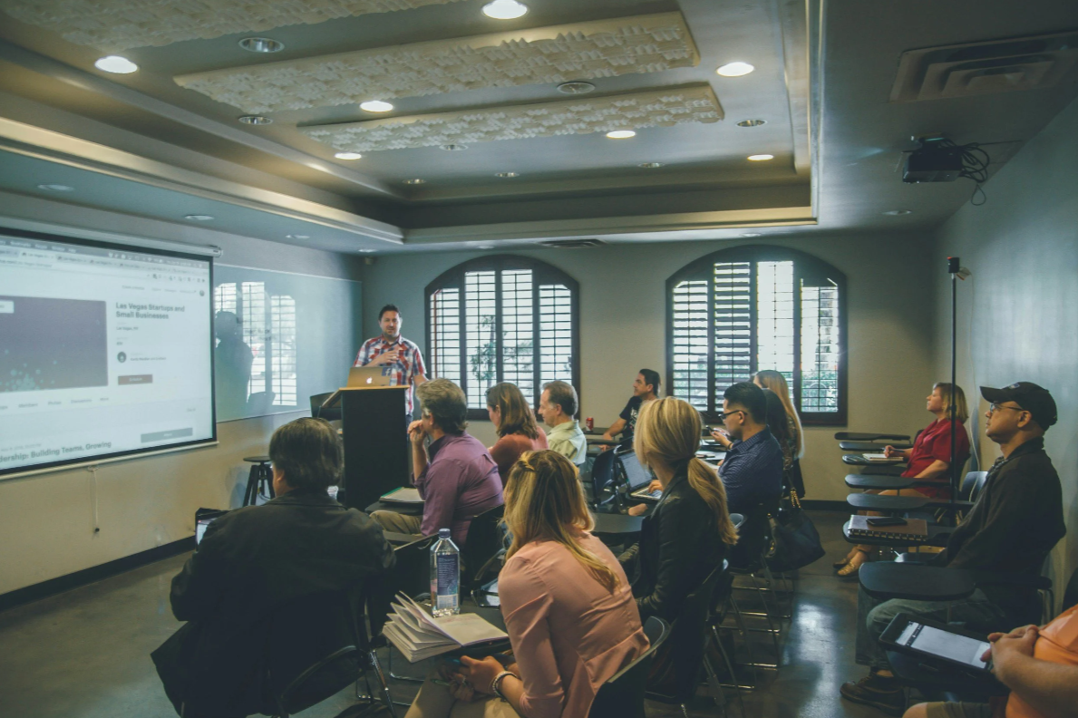 A group of professionals attend a workshop while a presenter speaks at the front of the room.