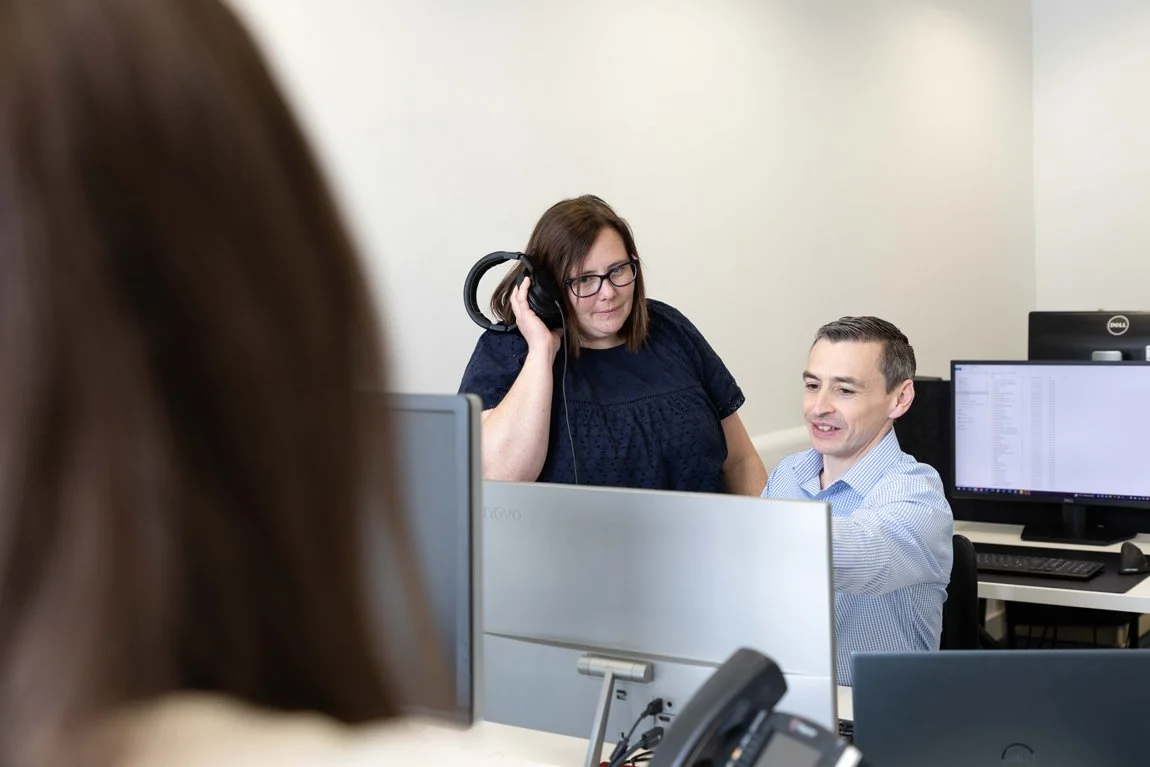 Office scene with three coworkers, one woman with glasses and headphones, two people visible in the background, computers, and office equipment.