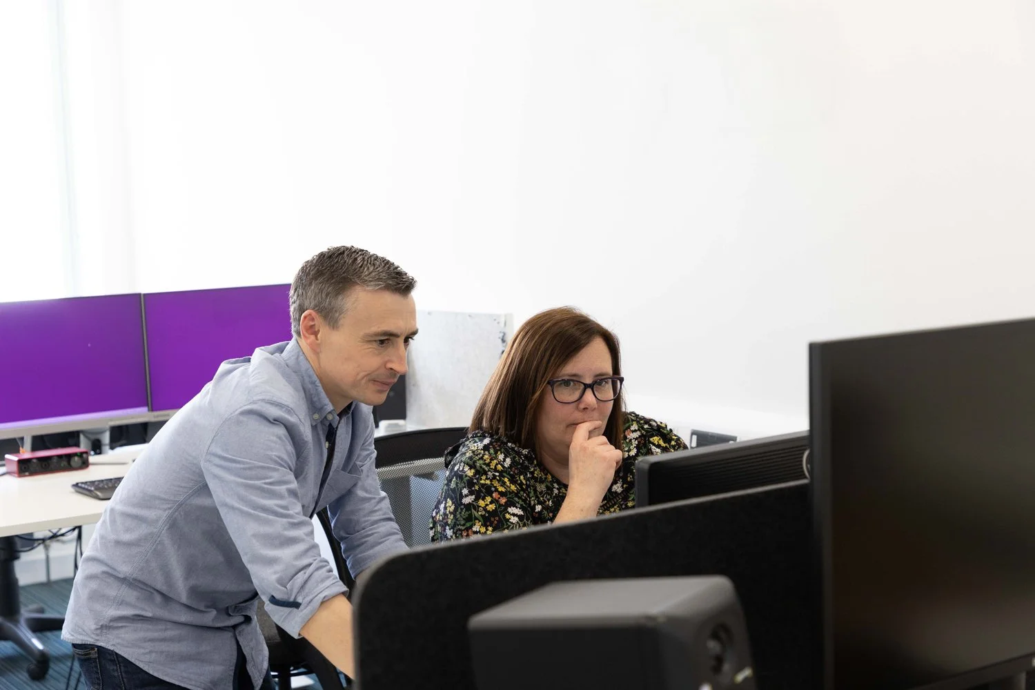 Two people working together at computers in an office, with one person standing and the other sitting, both focused on the screens.