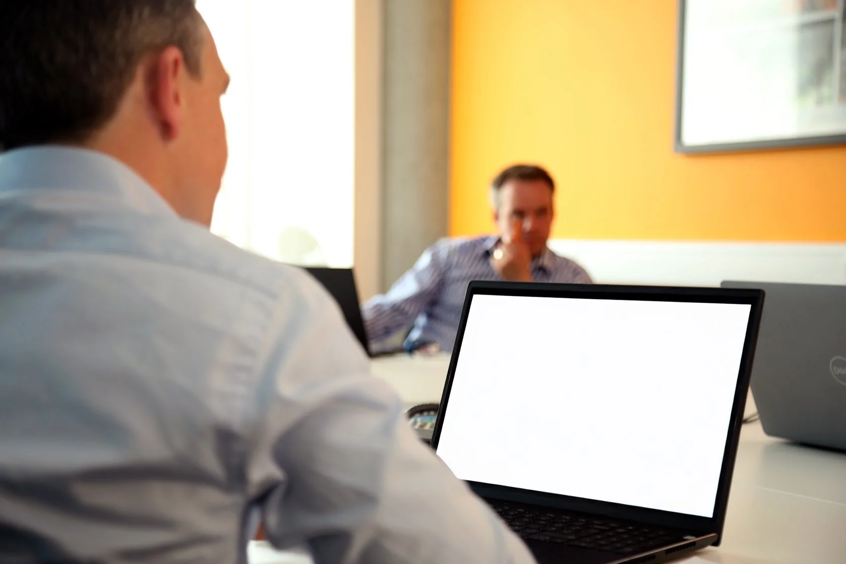 Two men in a meeting room, one using a laptop with a blank white screen, the other sitting in the background, talking, in front of a bright orange wall.