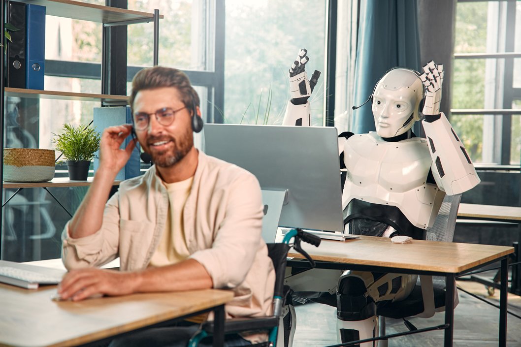 A man wearing glasses and a beige shirt sitting at a desk with a headset on, smiling. Behind him, a humanoid robot with a white body and black accents, wearing a headset, is sitting at another desk with its hands on its head. The room has large windows with blue curtains, a shelving unit, and a potted plant.