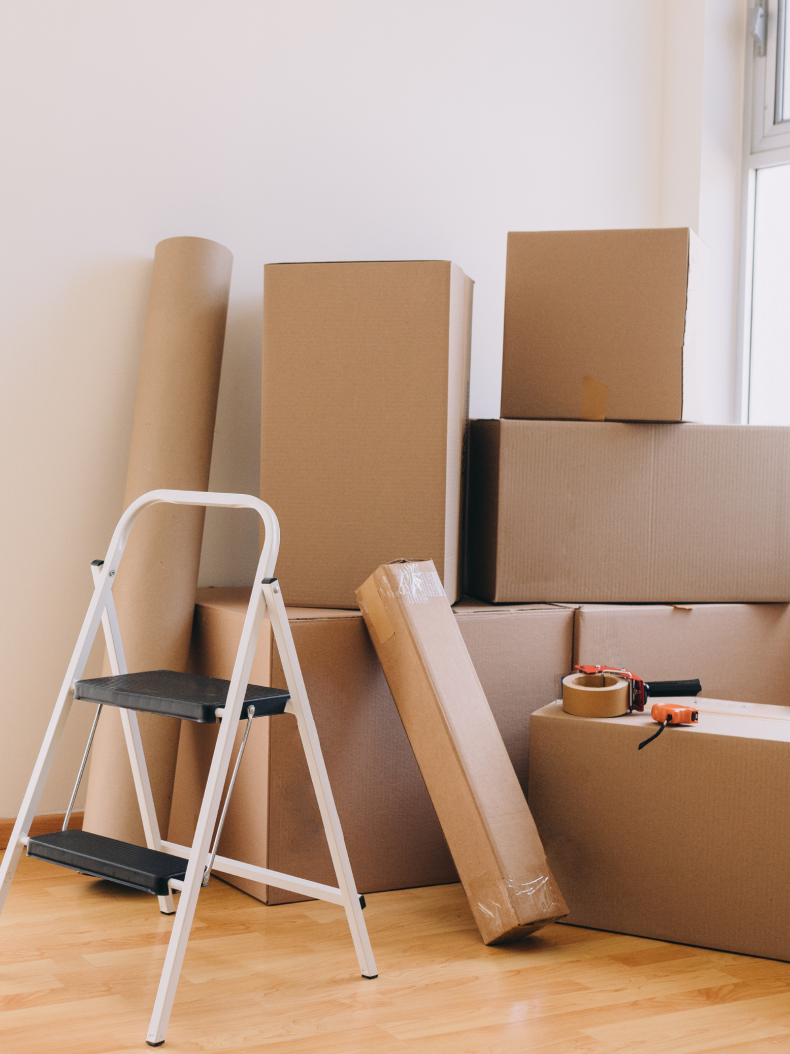 Stack of moving boxes and packing supplies inside a room, with a small black and white step ladder and a roll of packing tape.