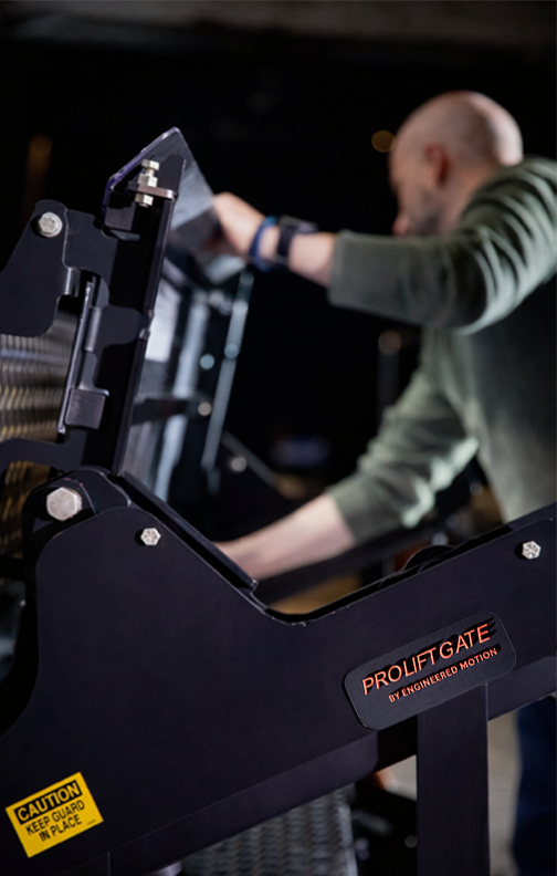 A person in a green shirt adjusting the ProLiftgate, with caution signs, in a dimly lit environment.