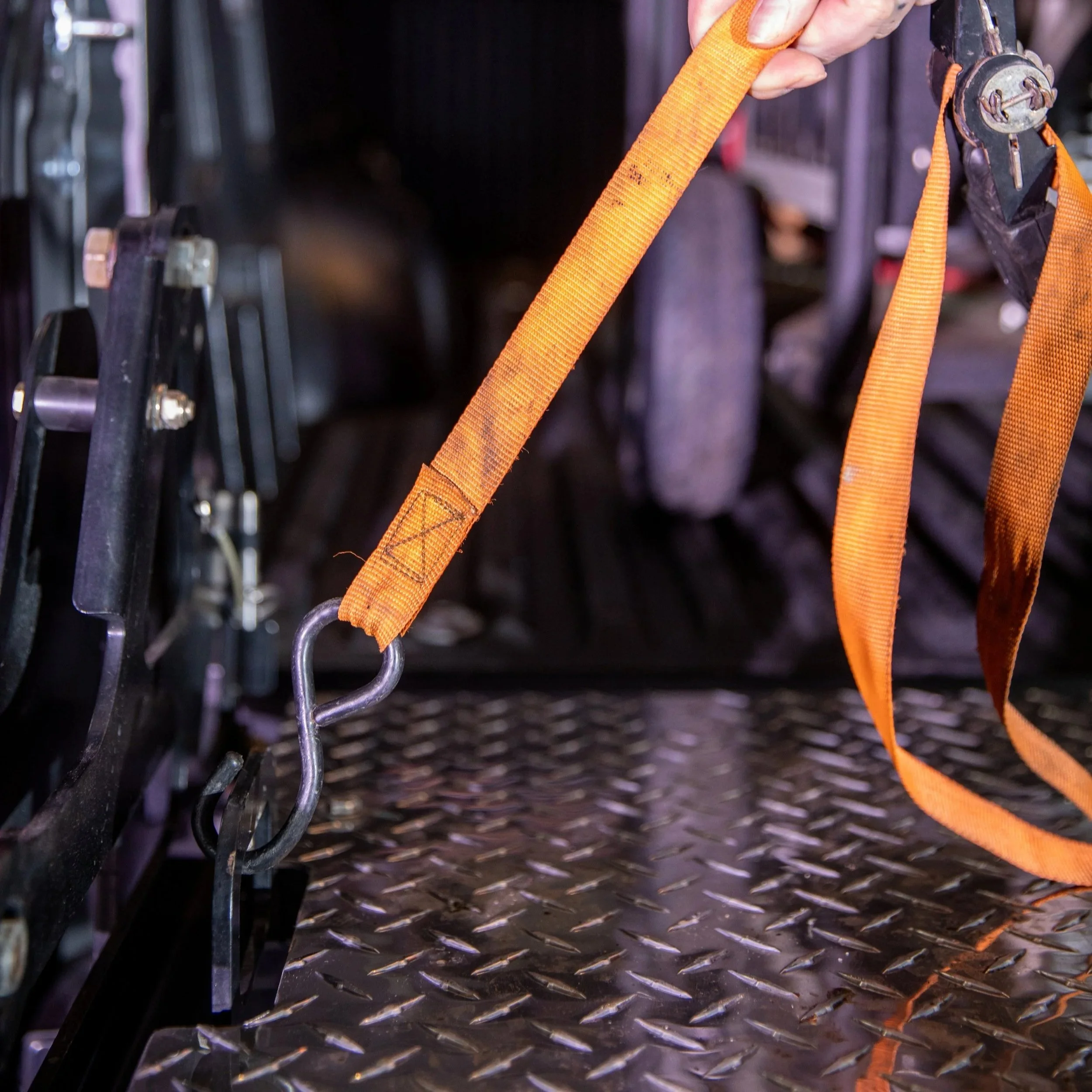 Close-up of an orange strap with a metal clasp attached to a harness system, on the ProLiftgate, with other hardware parts visible in the background.