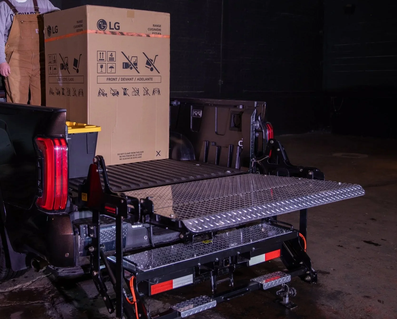 A pickup truck with an open tailgate and a black metal wheelchair lift platform. A large cardboard box is on the ProLiftgate, with printed instructions and icons for handling. The background is dark, likely an indoor or underground parking area.