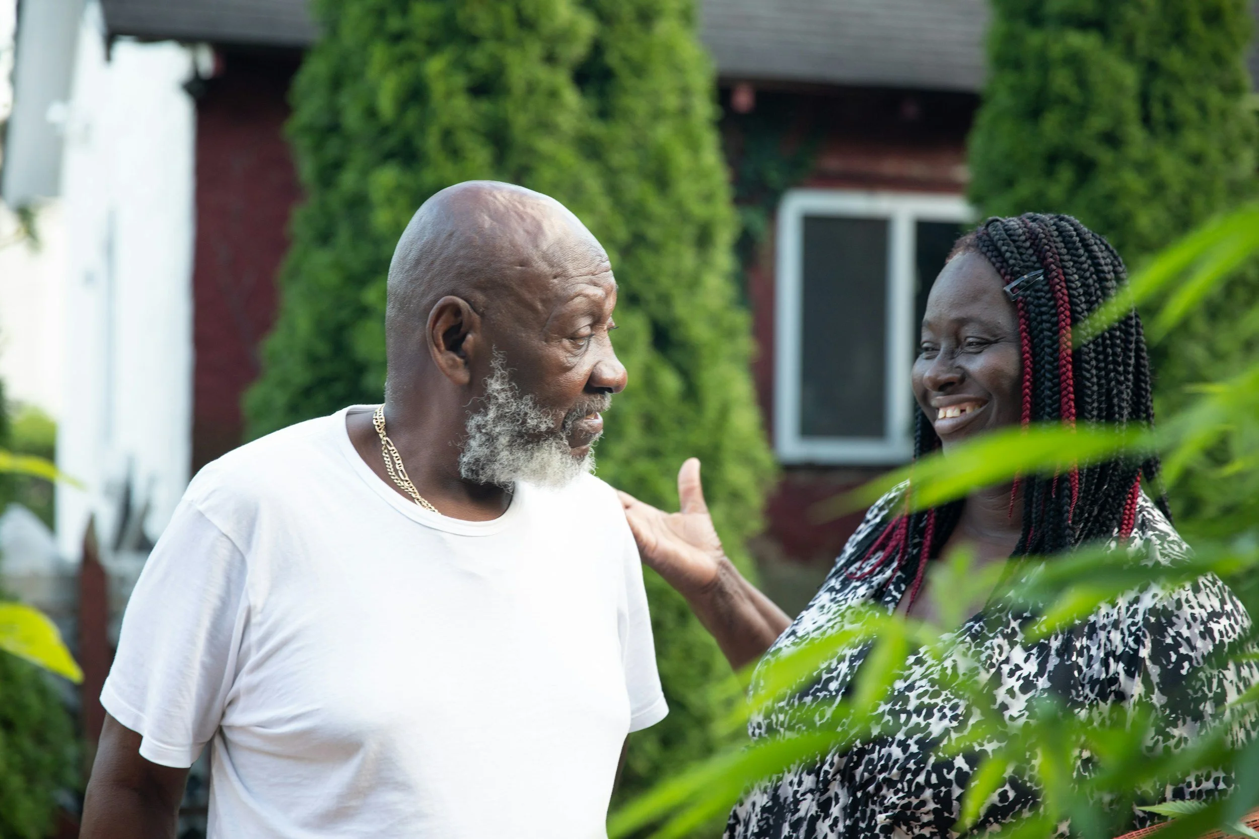 An elderly man with a gray beard and a white t-shirt smiling and talking to a smiling woman with braided hair in a garden with green plants and a house in the background.