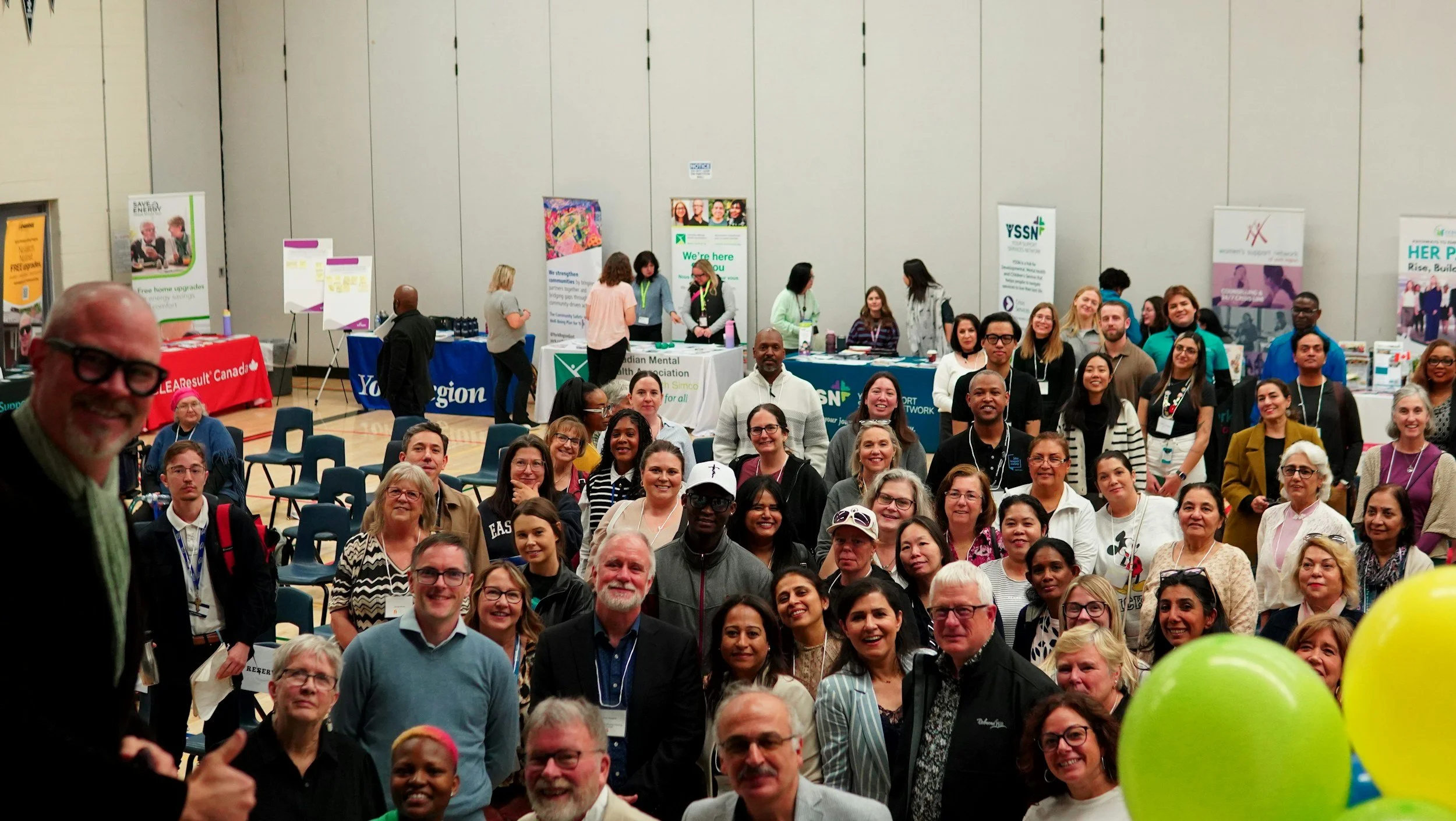 A large group of diverse people at a conference or event, posing for a photo in a hall with booths and banners in the background, some smiling and others looking at the camera.