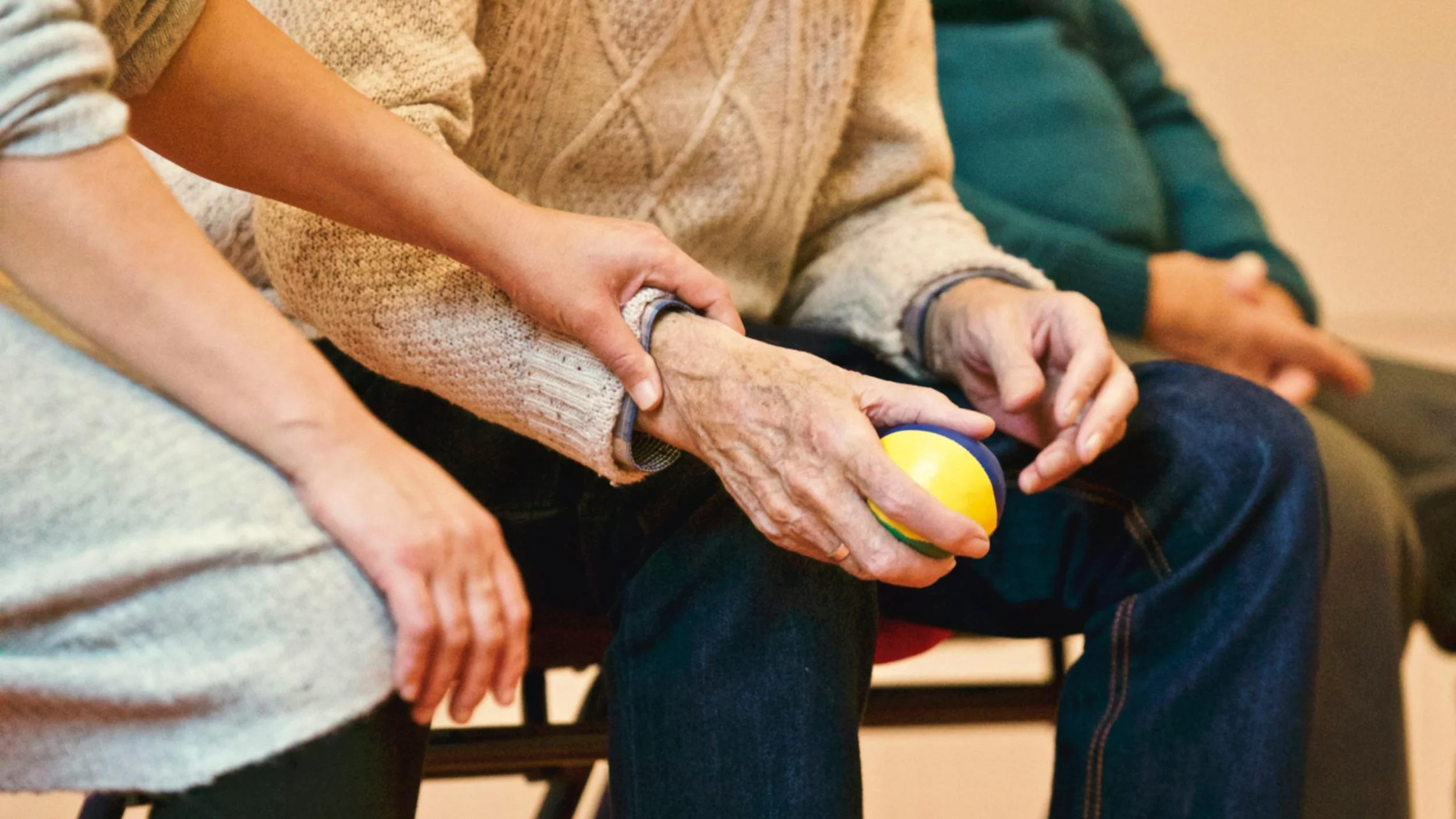 A close-up of an elderly person's hand holding a small yellow ball with a green stripe, with assistance from a caregiver or another person holding the elderly person's arm, in a social or medical setting.
