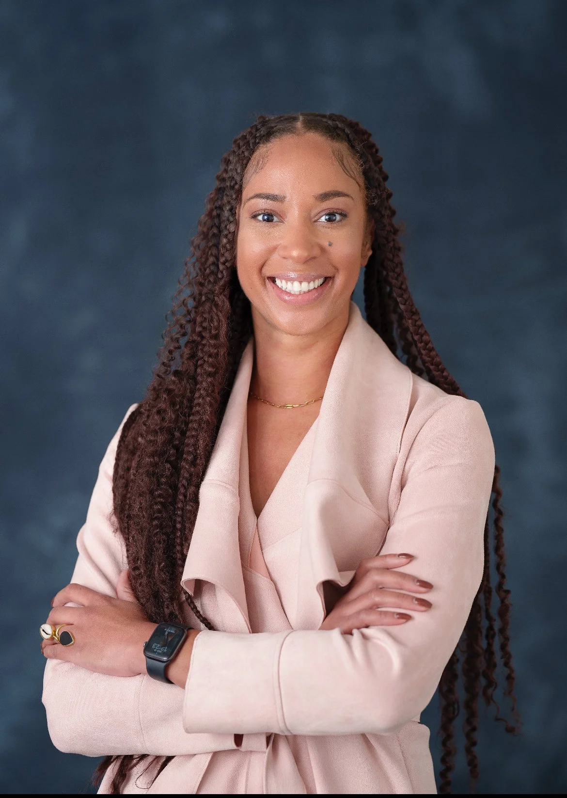 Professional woman with long braided hair smiling, wearing a light pink blazer, necklace, rings, and a smartwatch, standing with arms crossed against a dark blue background.
