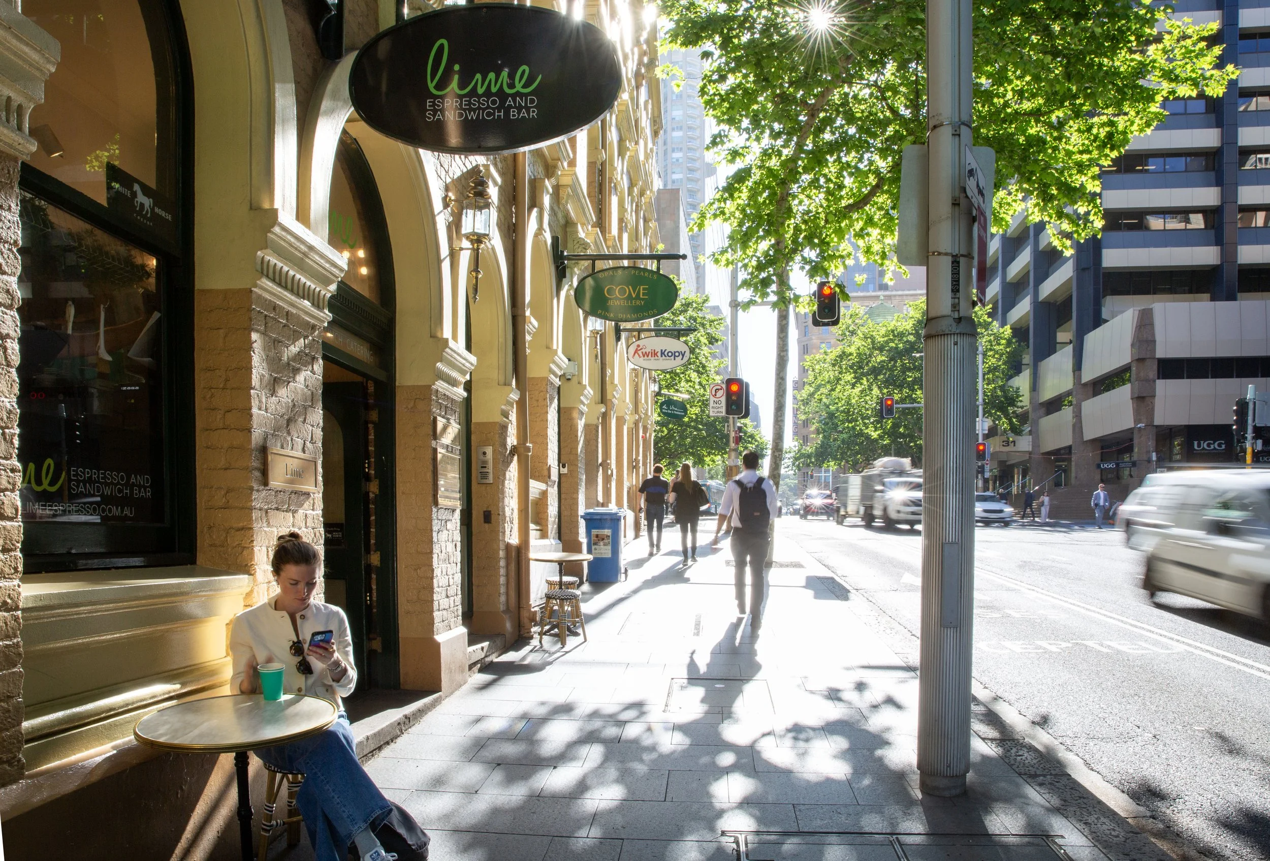 City sidewalk with pedestrians, cars on the street, trees, and storefronts including a cafe called 'Lime Espresso and Sandwich Bar' on a sunny day