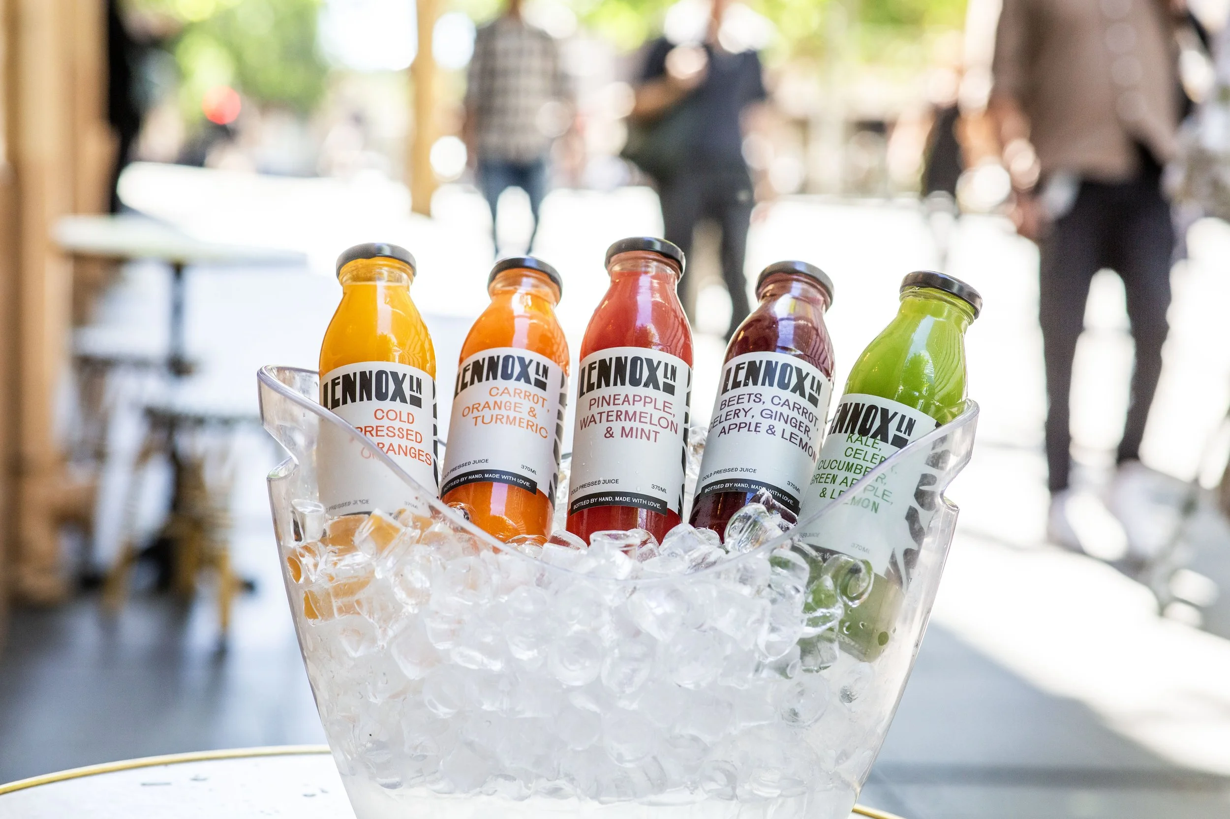 Assorted bottles of cold pressed juices in a glass container filled with ice on a table, with people in the background