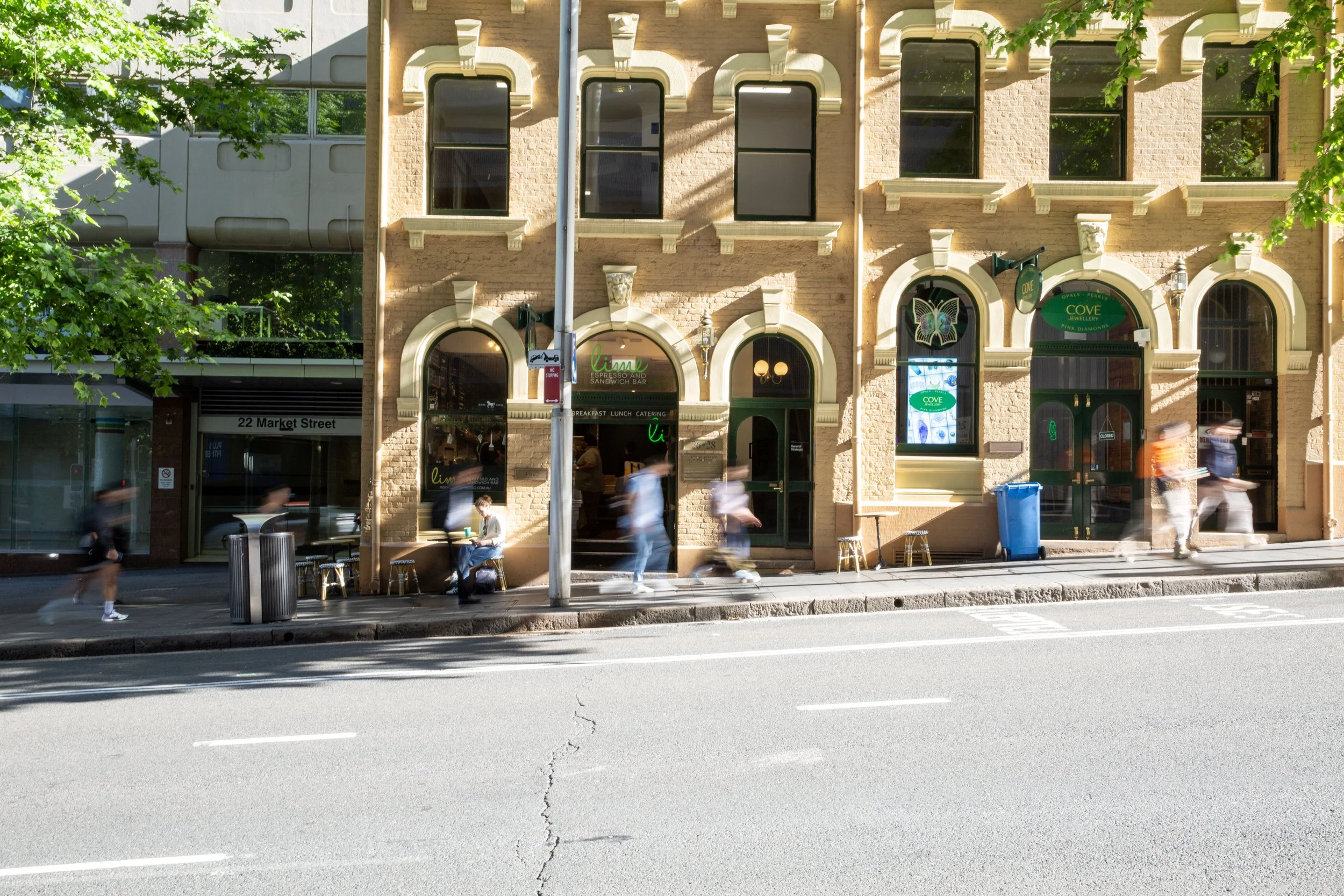 Street scene in front of a bakery or cafe called Lime with outdoor seating, people walking by, some sitting outside, a blue recycling bin, and a building with arched windows and a butterfly sign.
