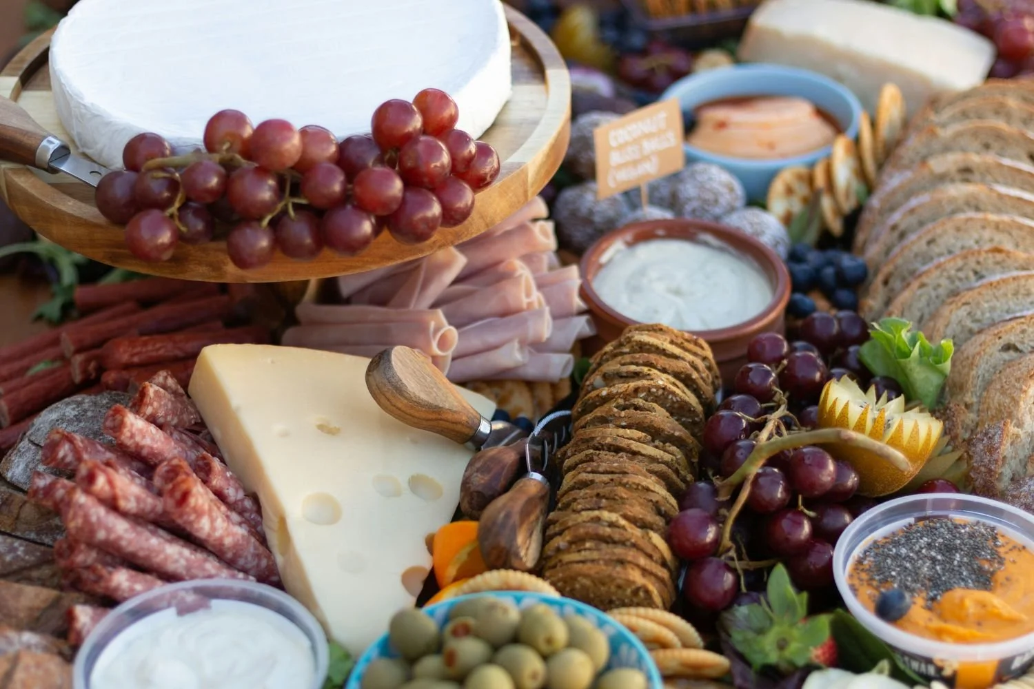 Assorted cheeses, cured meats, crackers, grapes, olives, dips, and charcuterie on a table.