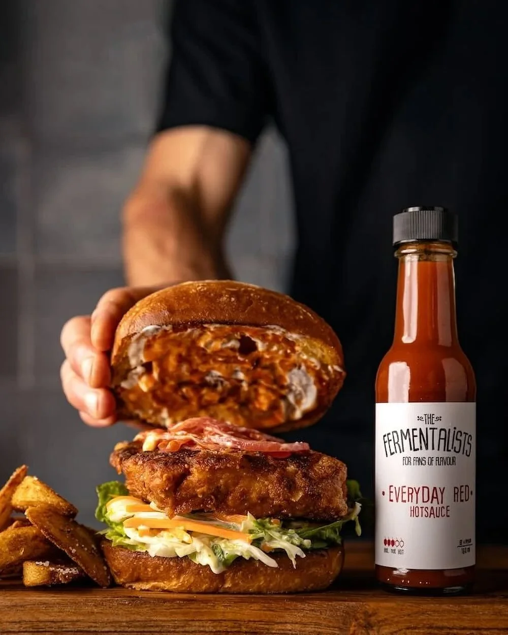 A person holding a half-eaten burger with a chicken patty, lettuce, tomato, and cheese on a bun, with a side of potato wedges and a bottle of hot sauce labeled "The Fermentalists Everyday Red Hot Sauce".