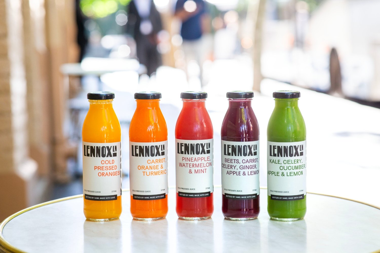 Five bottles of cold pressed juice with various fruit and vegetable flavors, arranged on a white surface in a row. The background is blurred with people and trees visible.