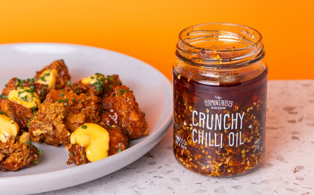 Fried chicken pieces with sauce on a white plate with a jar of crunchy chili oil on an orange background.