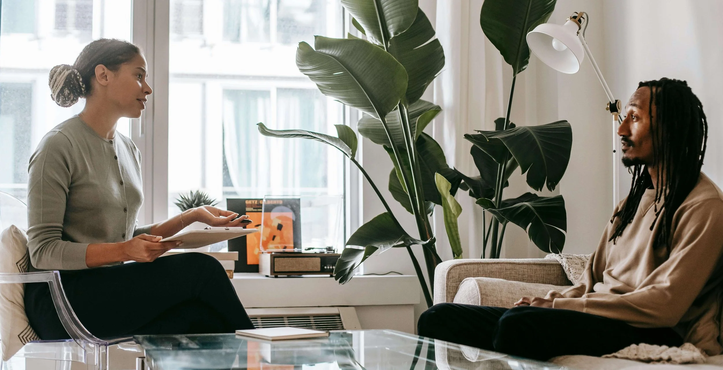 Therapist holding a clipboard while talking with an adult client in a modern counseling office with natural light and greenery.