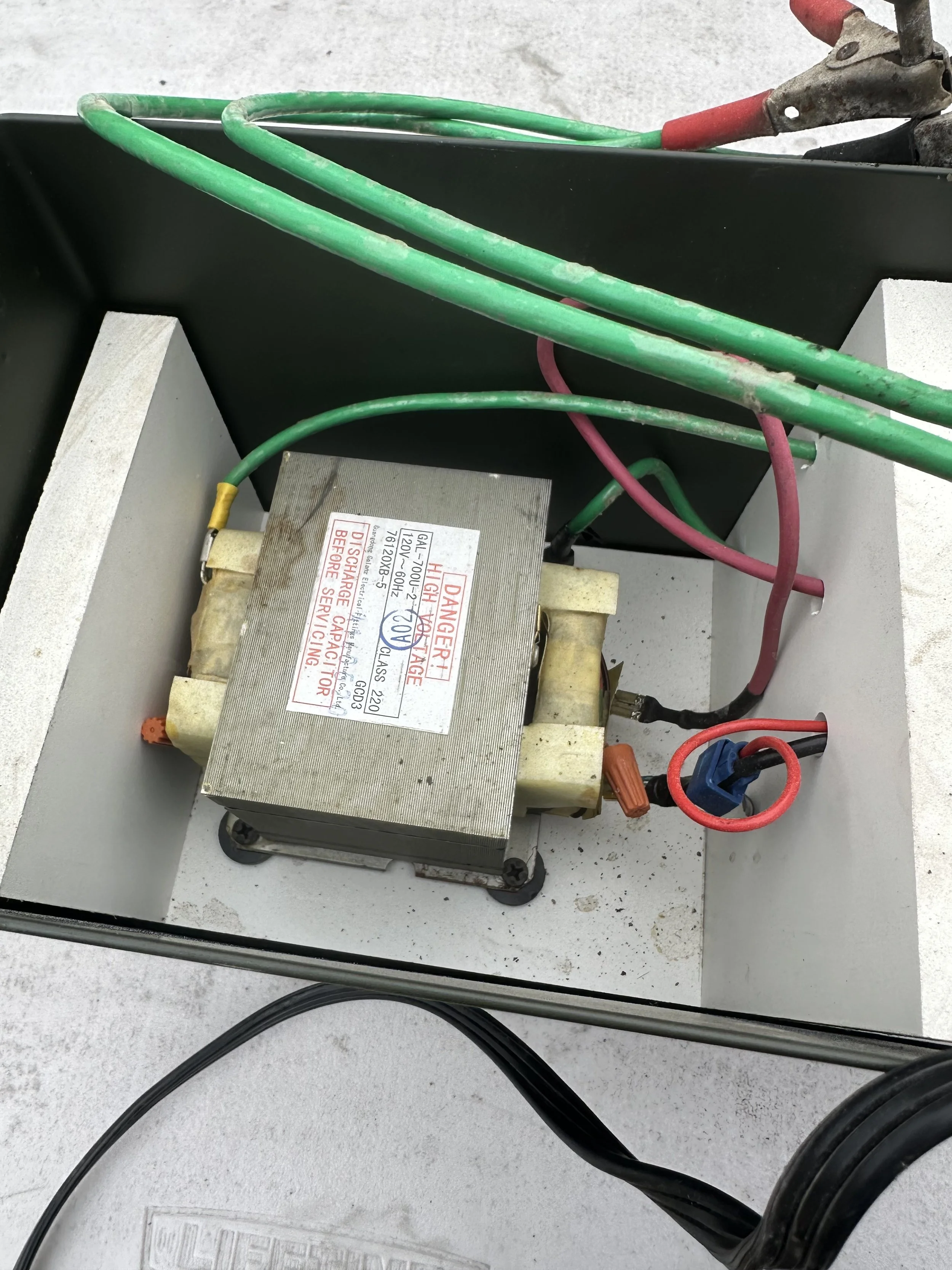 Electrical transformer with colored wires connected to it, housed in a gray metal box, mounted on a light gray surface.