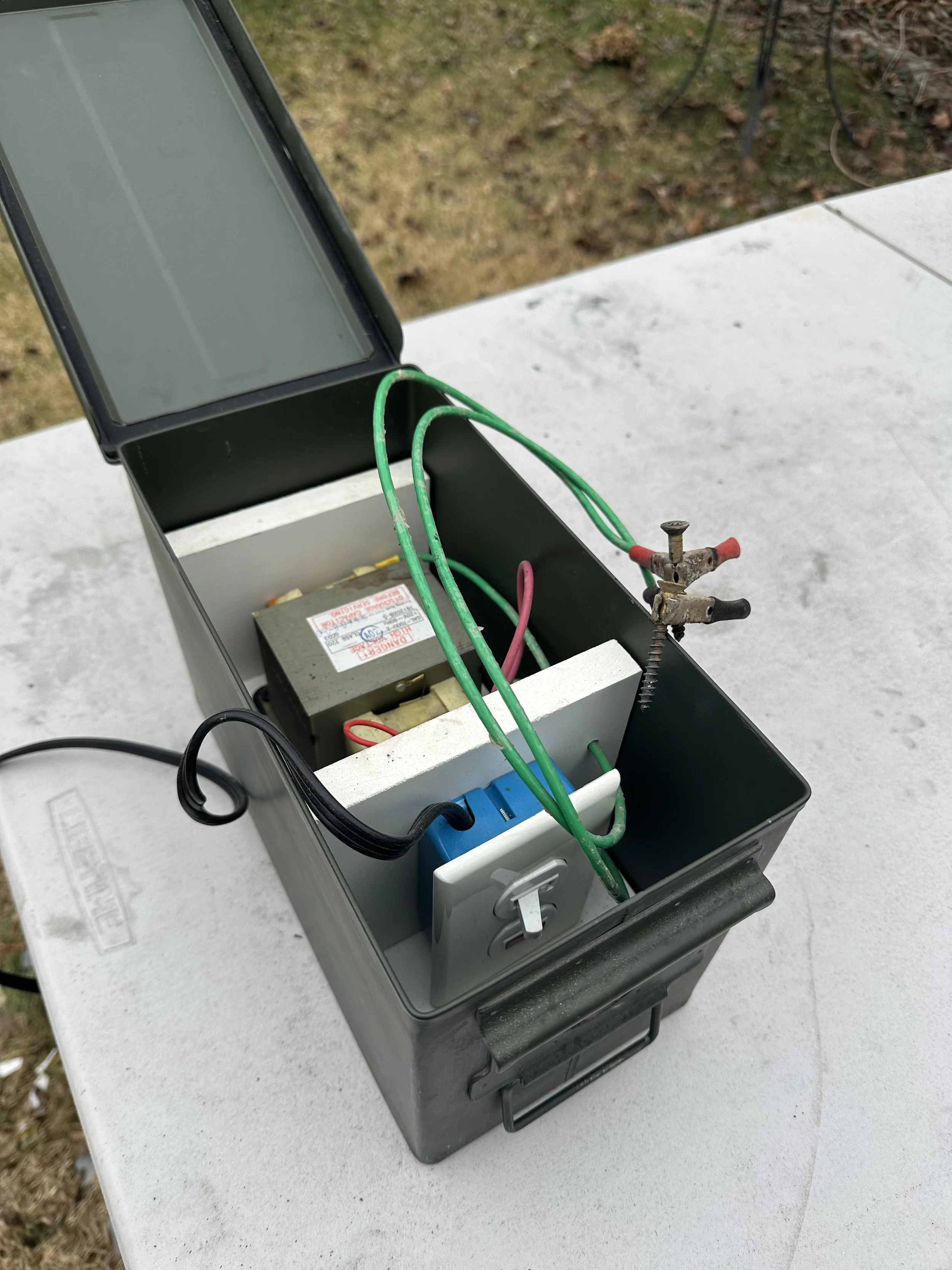 A metal box containing electrical components, wires, and terminals on a white table outdoors.