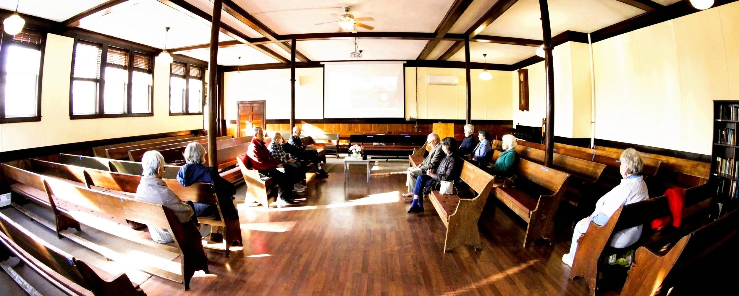 A group of people attending a presentation or discussion in a room with a large projection screen. The screen shows an image of an older man with a beard. The room has wooden walls, a wall clock, and several flower arrangements on tables.