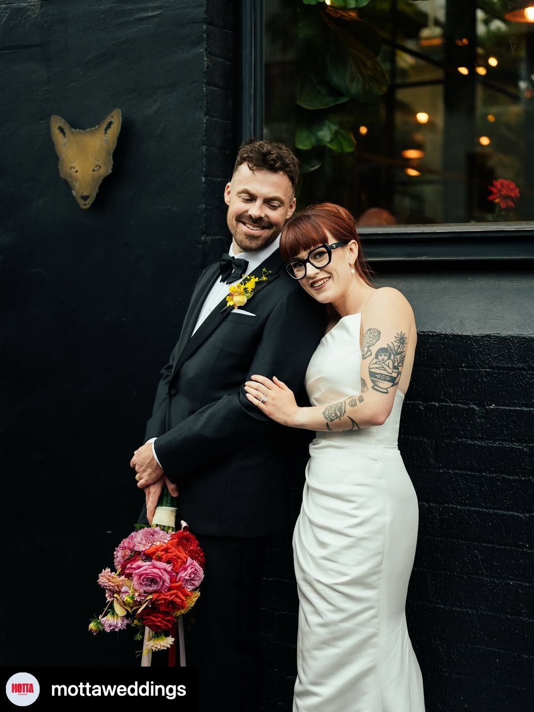 A newlywed couple standing close together, smiling, with the bride leaning on the groom's shoulder. The groom is wearing a tuxedo with a yellow floral boutonniere and holding a bouquet of colorful flowers. The bride is wearing a white satin wedding dress with tattoos visible on her arms, and black-rimmed glasses. They are in front of a black wall with a fox head wall decor, and a window with plants and lights inside.