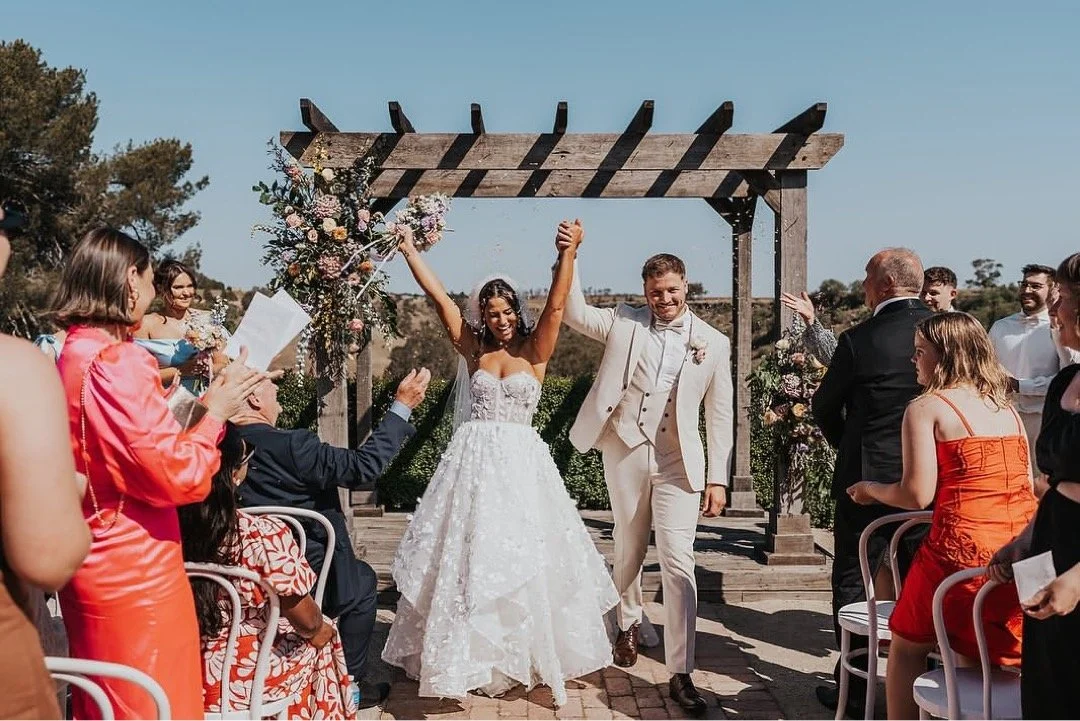 A newlywed couple walking down the aisle, holding hands and raising their arms in celebration, surrounded by wedding guests during an outdoor wedding ceremony.