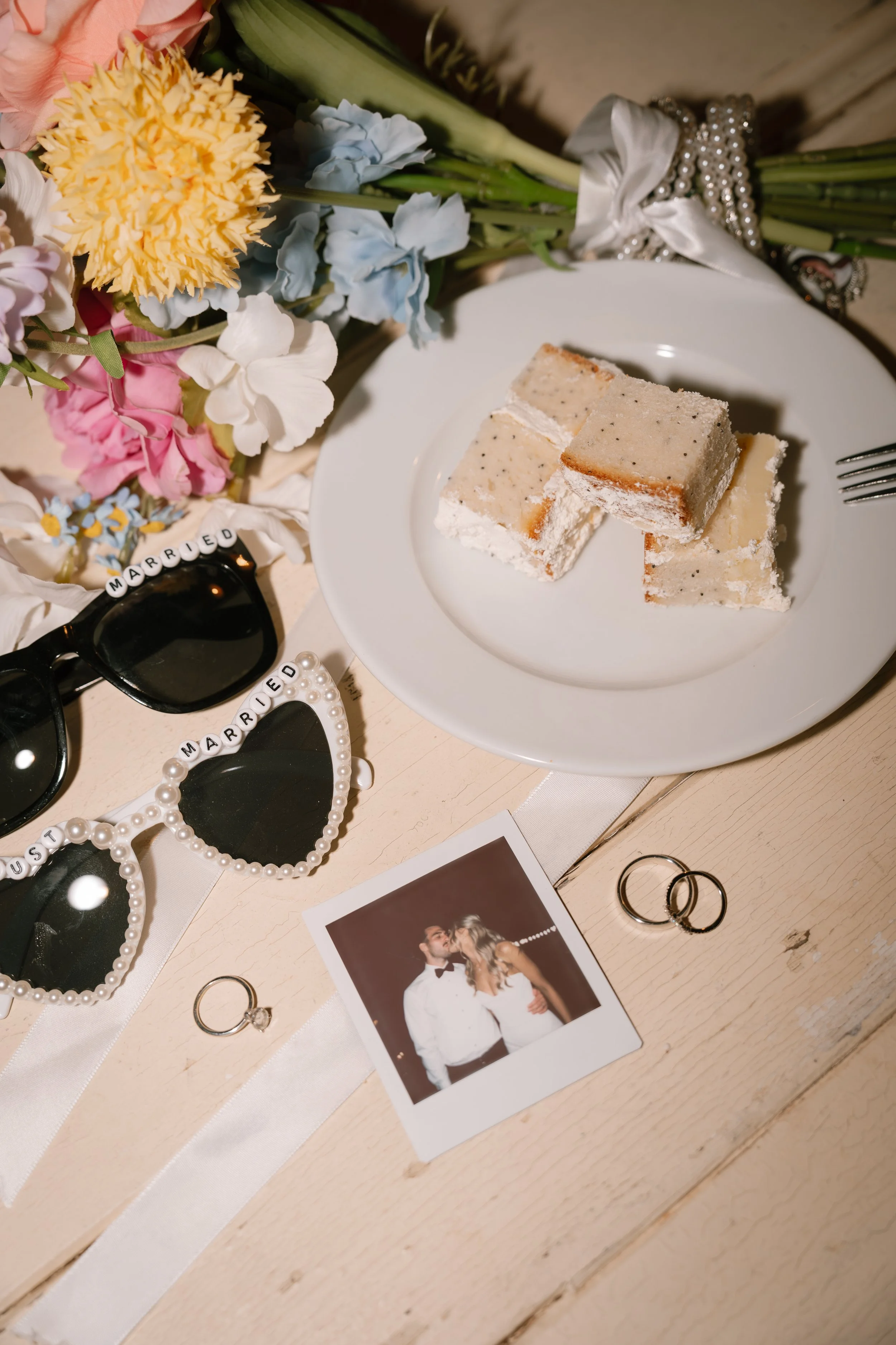 A picnic table with a white plate holding five pieces of almond-flavored cake, surrounded by wedding-related items such as a photo of a bride and groom, wedding rings, pearl sunglasses, and a bouquet of mixed flowers. The setting appears celebratory and romantic with wedding accessories.