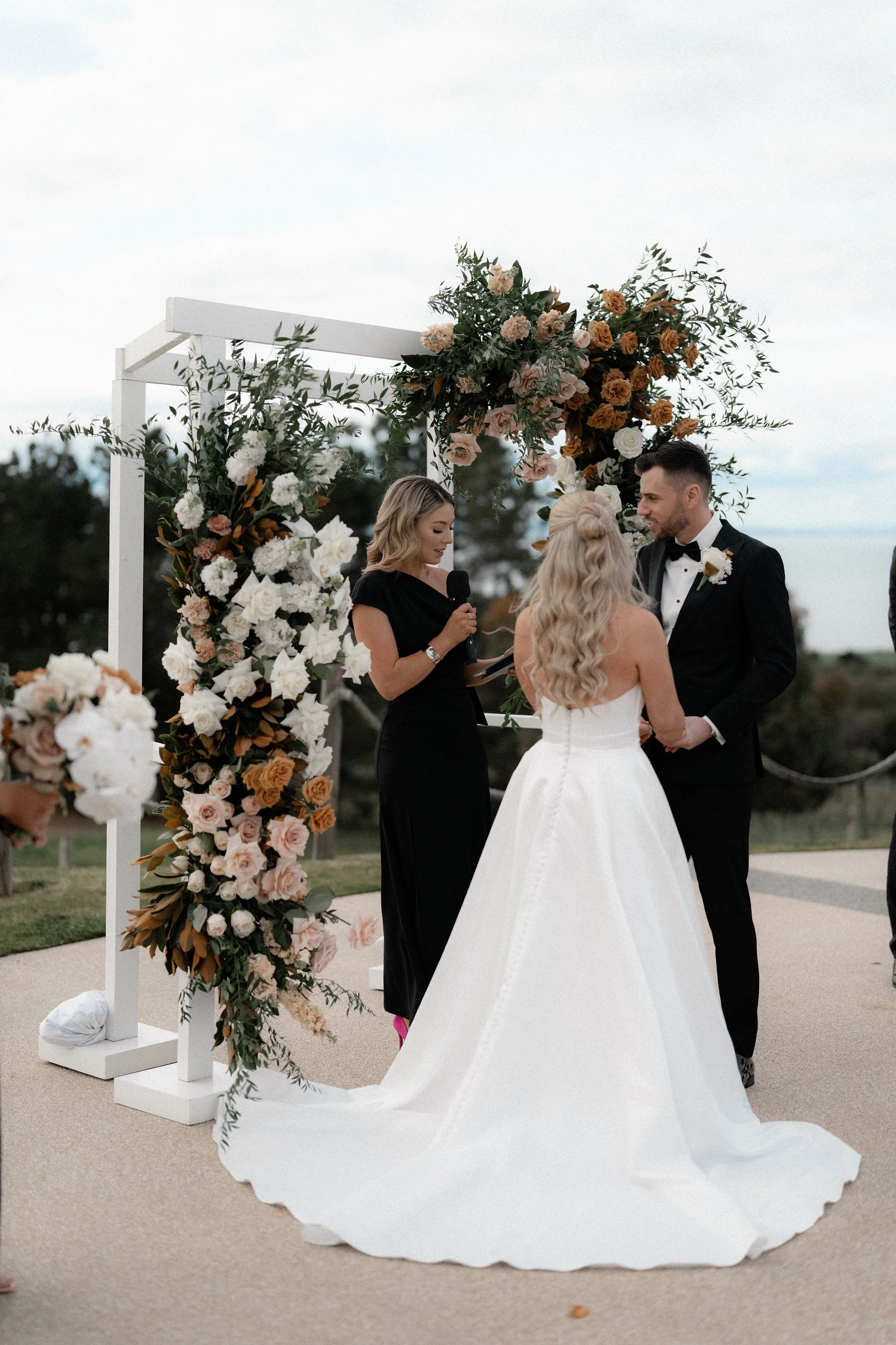 A couple getting married, holding hands, with a female officiant holding a microphone. They stand under a floral archway overlooking an outdoor landscape.