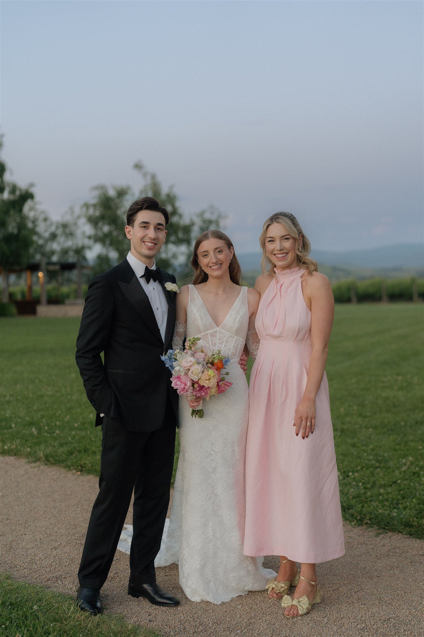 A wedding party with a groom in a black tuxedo, a bride in a white lace dress holding a colorful bouquet, and a woman in a pink dress, standing outdoors on a grassy area with trees and rolling hills in the background during the evening.
