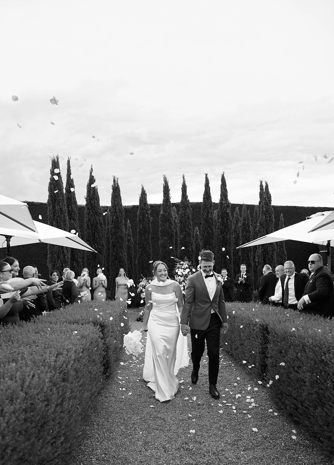 A black and white photo of a bride and groom walking down an outdoor aisle, surrounded by guests throwing flower petals. The bride is in a long white dress holding a bouquet, and the groom is in a suit. There are tall trees and umbrellas in the background.