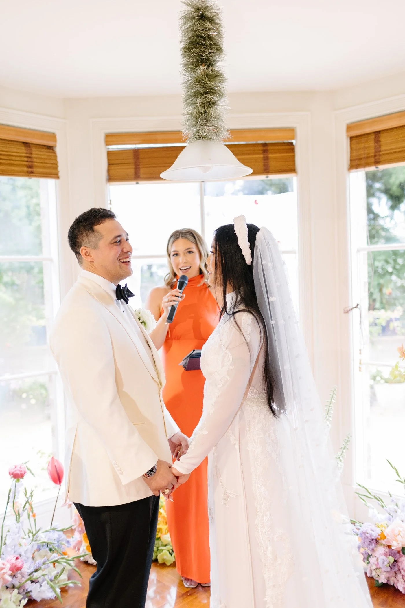 Bride and groom holding hands during their wedding ceremony, with officiant speaking into a microphone inside a brightly lit room with large windows and floral decorations.