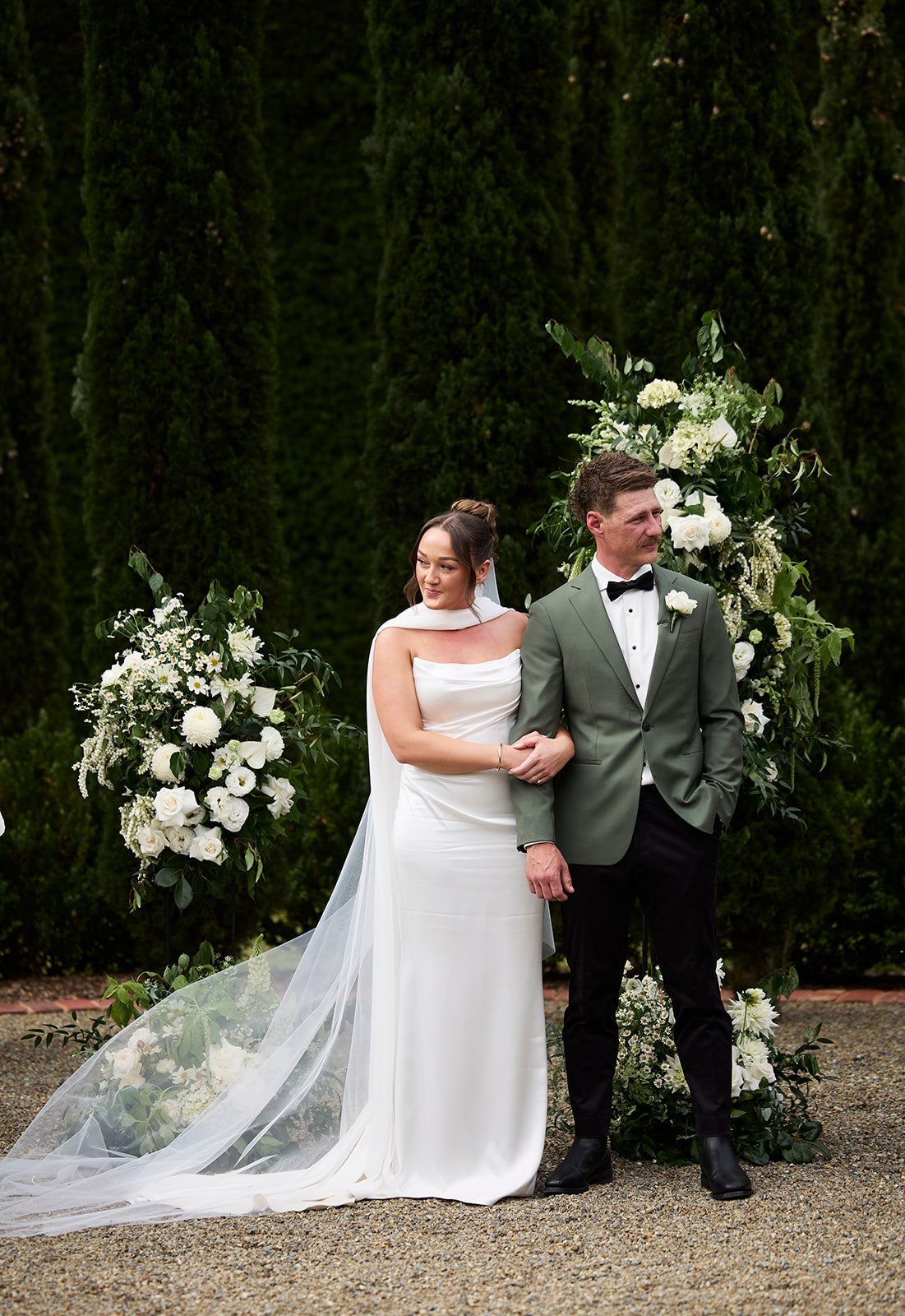 A bride and groom at their wedding ceremony standing in front of a floral arch with white flowers and greenery. The bride is wearing a white wedding gown with a veil, and the groom is in a green suit with a black bow tie.