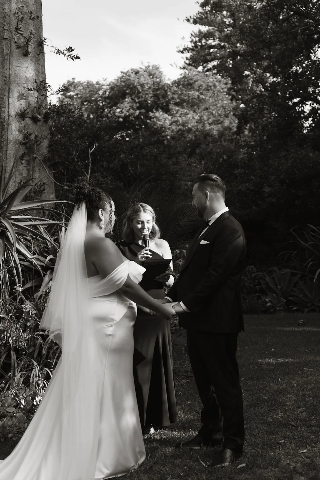A wedding ceremony outdoors with a bride and groom holding hands, standing in front of an officiant who is reading from a book, surrounded by trees and foliage.