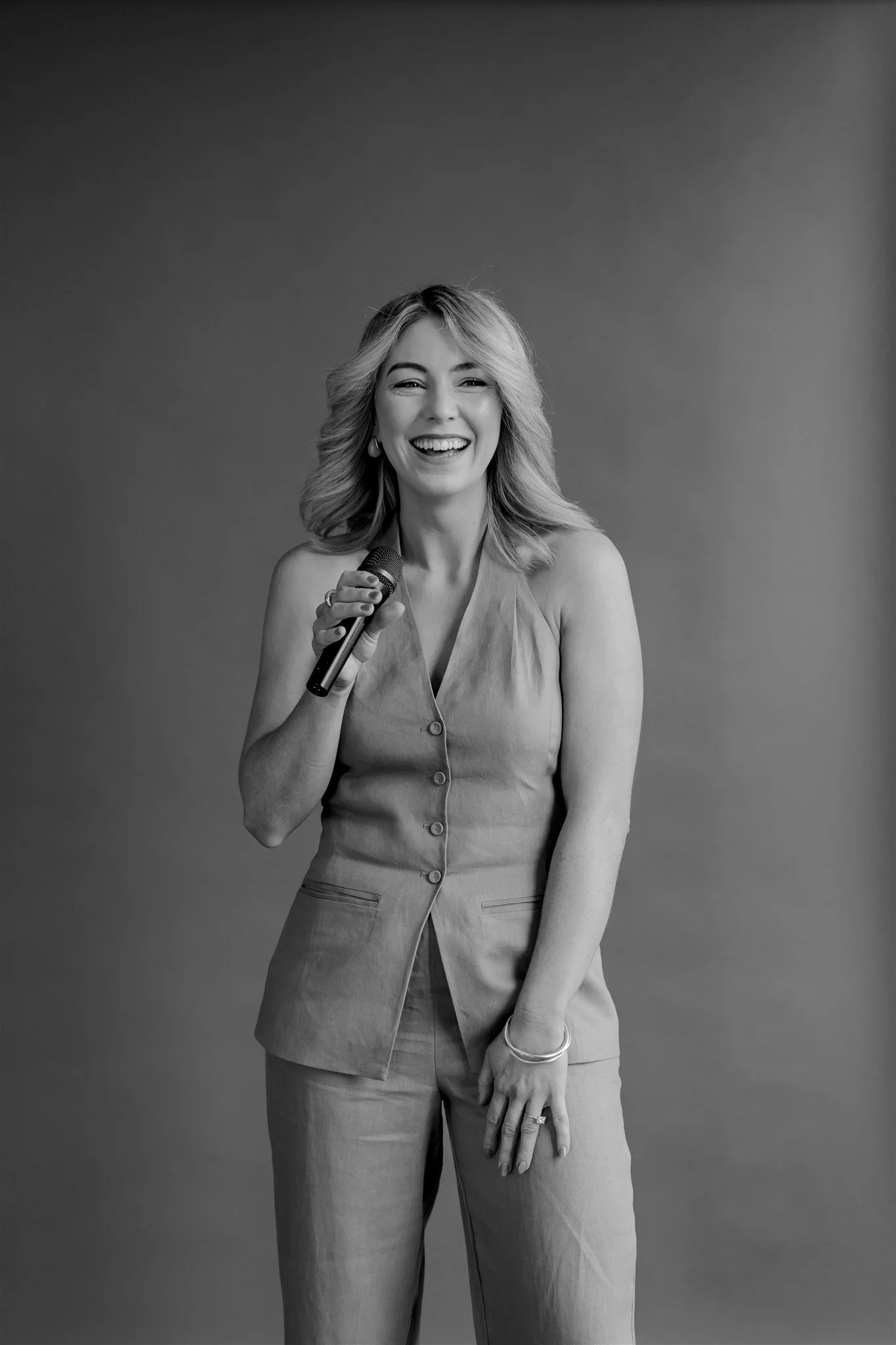 A woman with wavy blonde hair, smiling, holding a microphone, wearing a sleeveless blazer and matching pants, standing against a plain background.