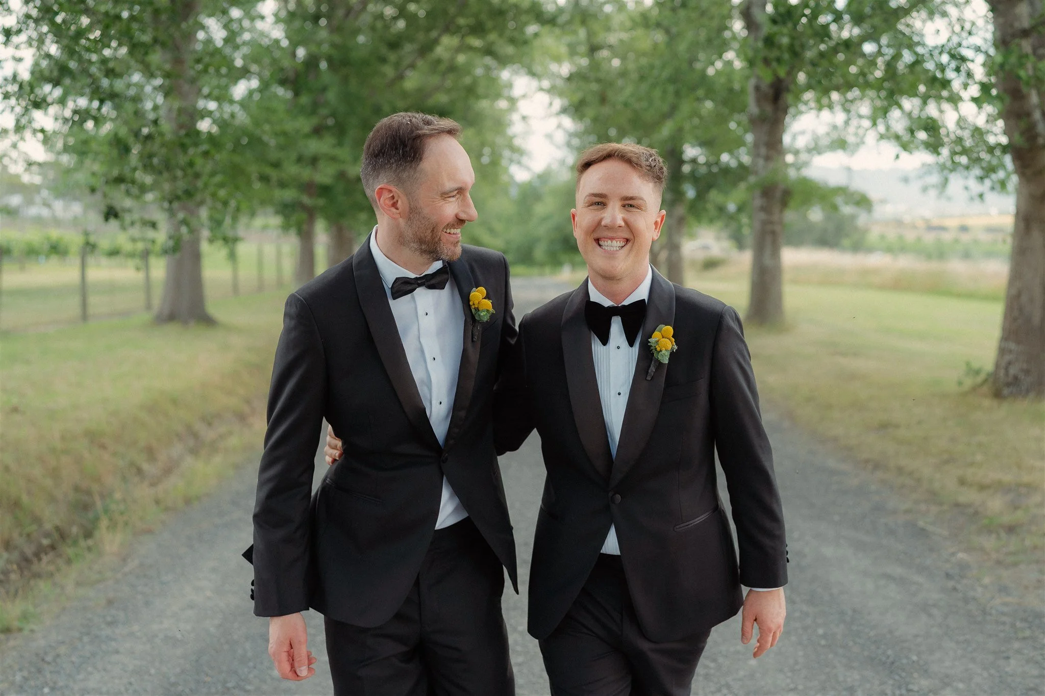 Modern house with wooden exterior in front of mountains, with a large window showing two people inside, surrounded by yellowish and green vegetation.