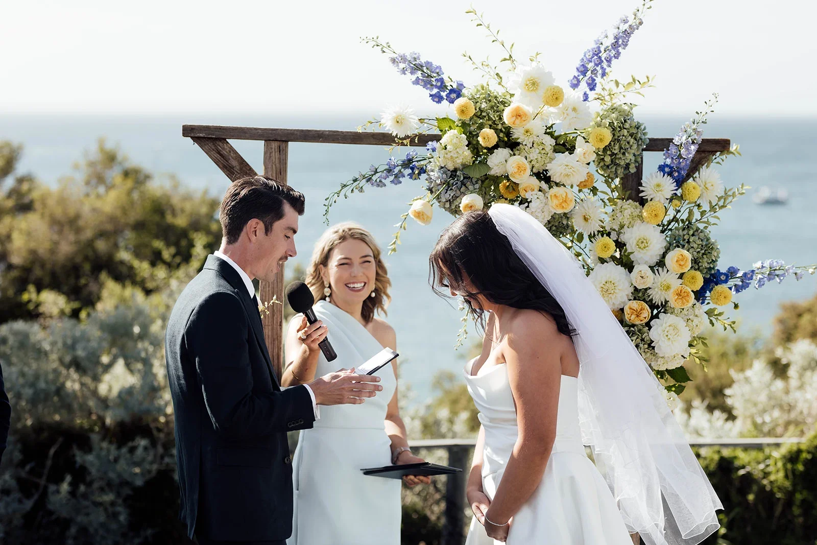 Bride and groom exchanging vows during outdoor wedding ceremony by the sea with officiant smiling in the background.