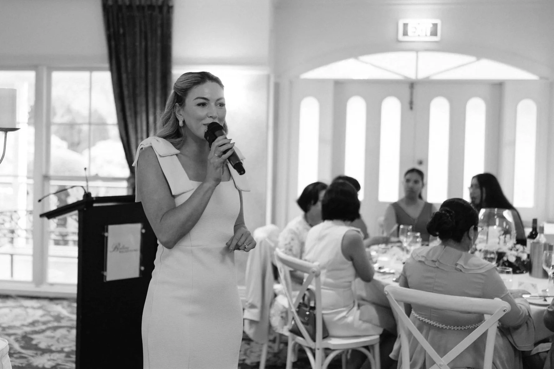 A woman speaking into a microphone at a formal event, with people seated around a dining table, in a room with large windows and an exit sign.