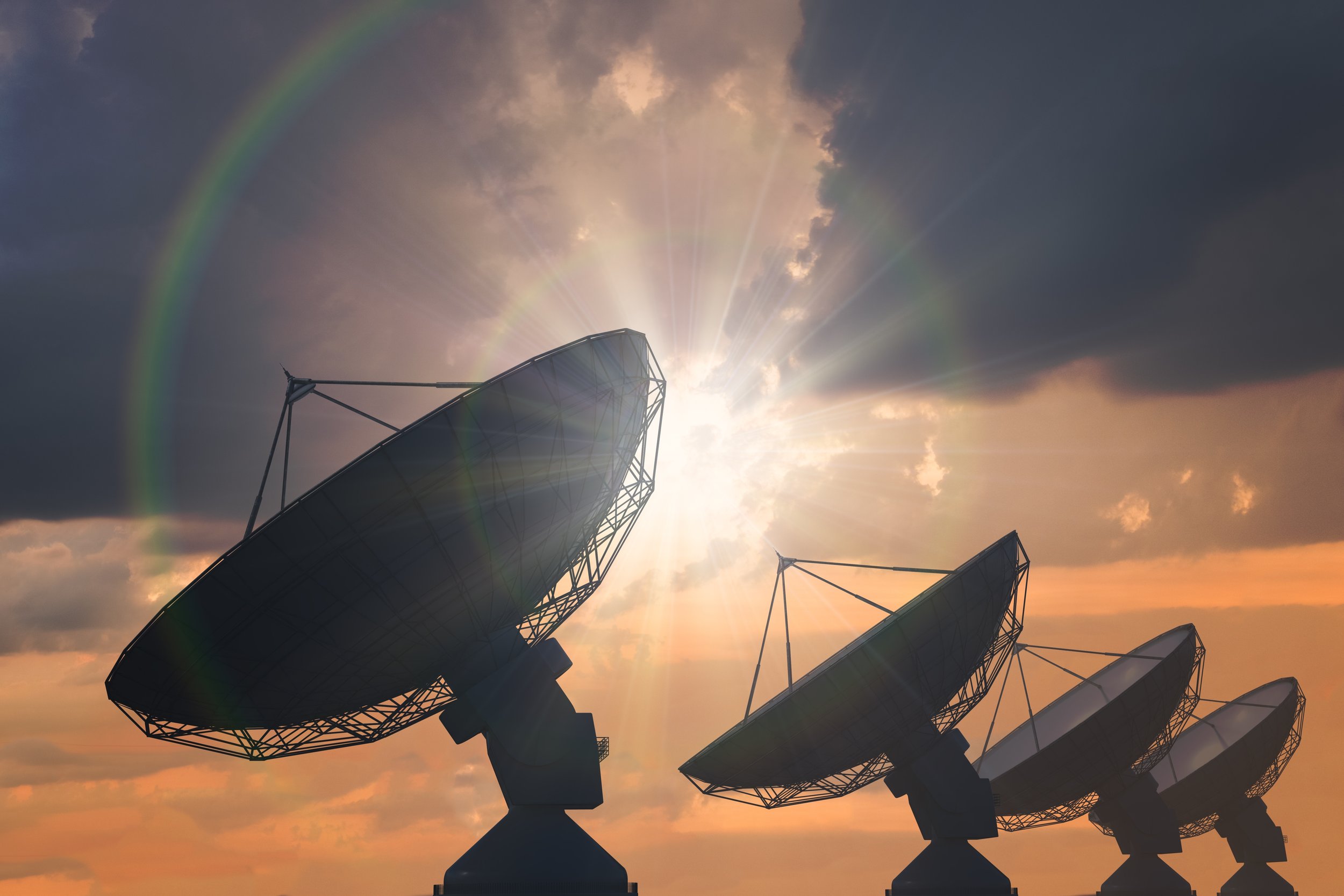 Four satellite dishes pointing skyward against a sunset sky with clouds and a rainbow haloing the sun, representing the vast reach of space exploration and the space industry.