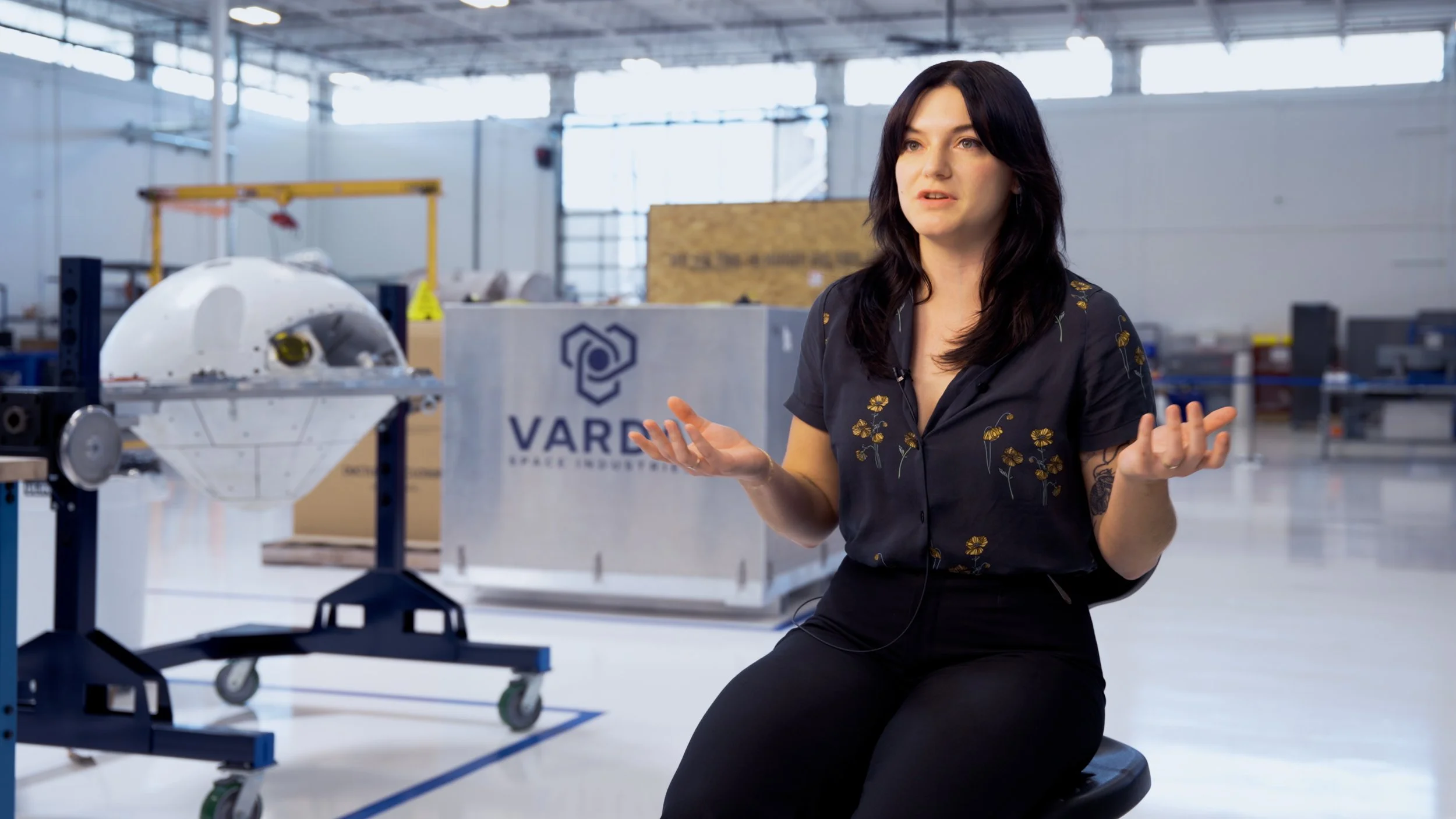 Space industry scientist speaking on camera during brand film production at a space manufacturing facility, with flight hardware in the background.
