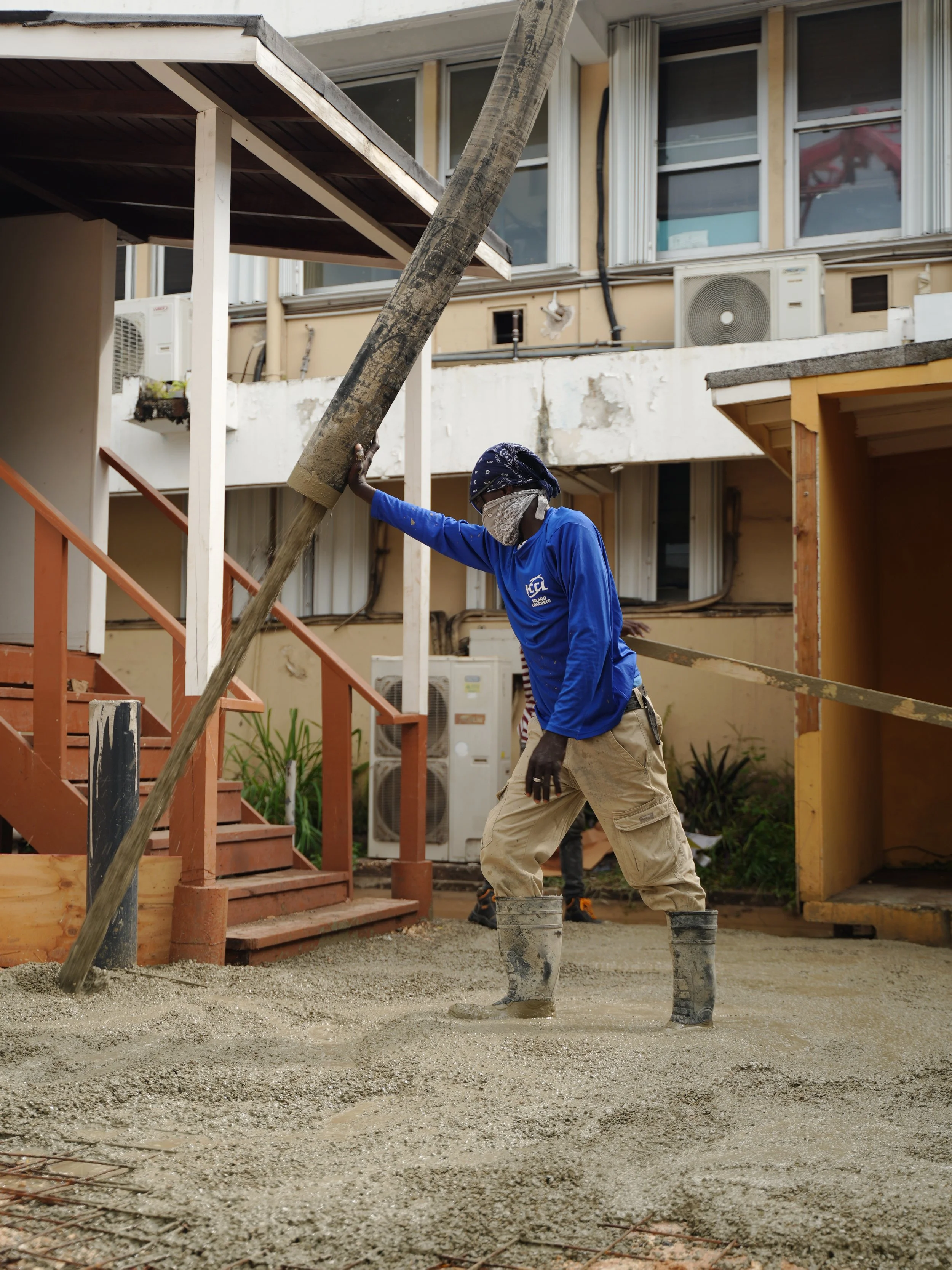 A construction worker pouring concrete on a building site, wearing boots, a blue long sleeve shirt, and a bandana head covering, with a building and stairs in the background.