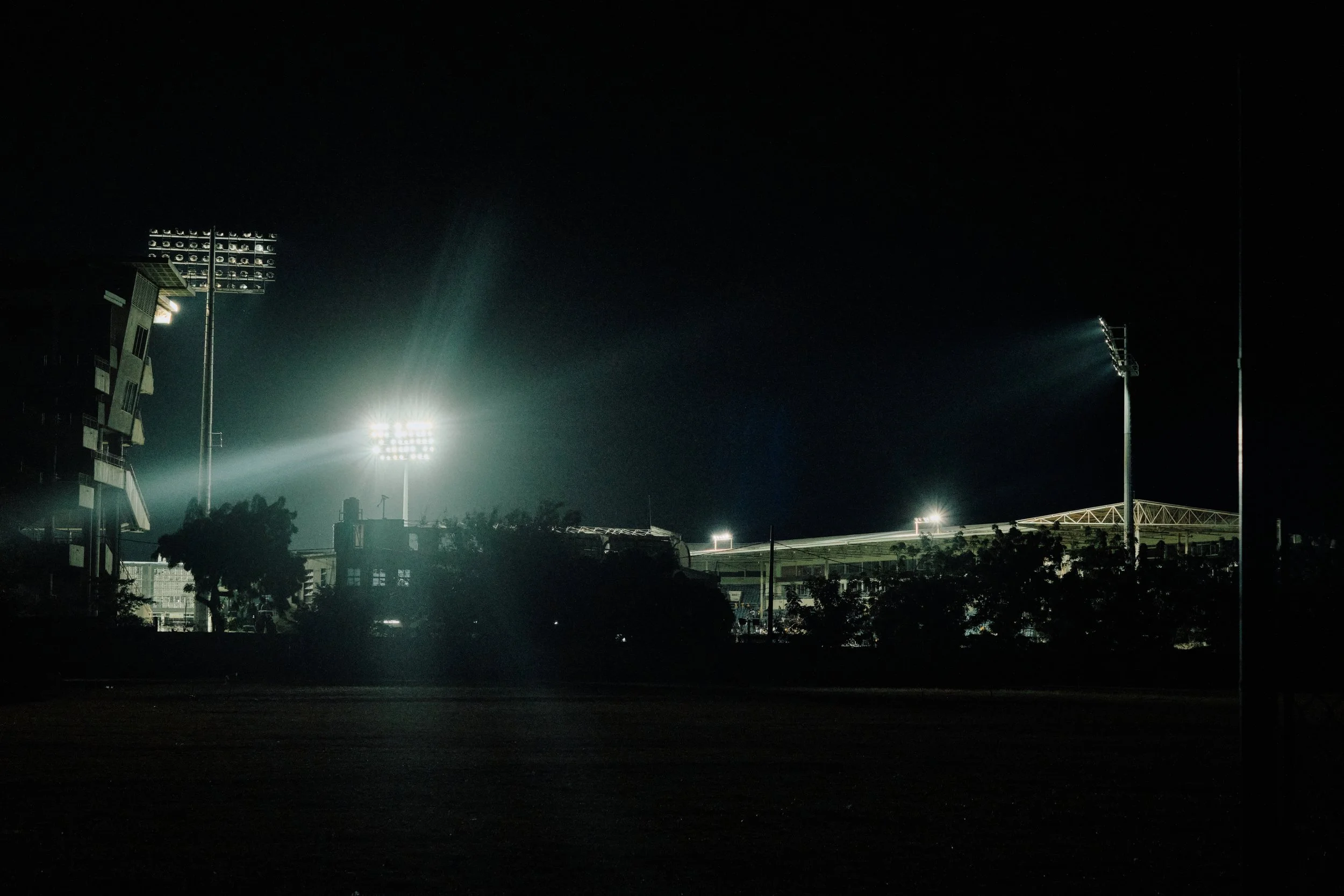 A sports stadium at night with bright floodlights illuminating the field and surrounding area, dark sky overhead, trees and structures in the foreground.