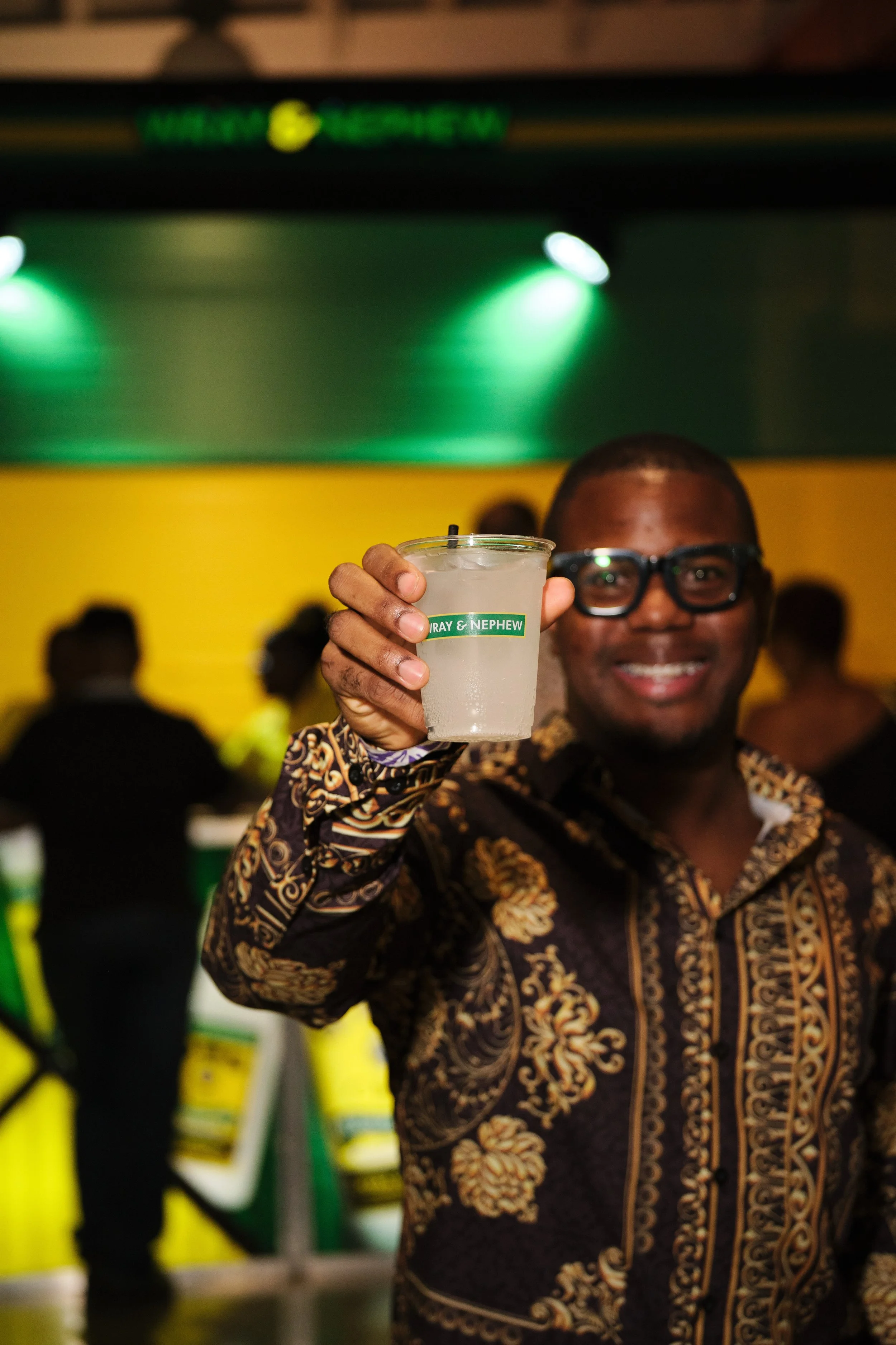 A man in a patterned shirt is smiling and holding up a clear drink in a plastic cup with a straw, in a lively indoor setting with yellow and green decor.