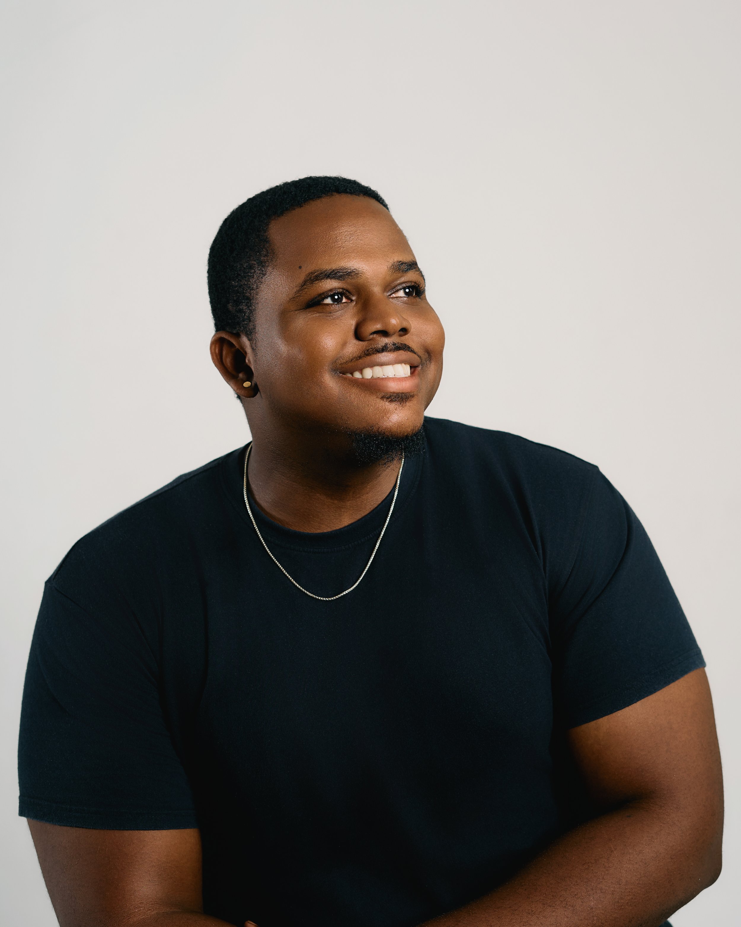 A smiling young person with dark skin, short black hair, wearing a black t-shirt, a silver necklace, small earrings, and looking to the side against a plain light background.