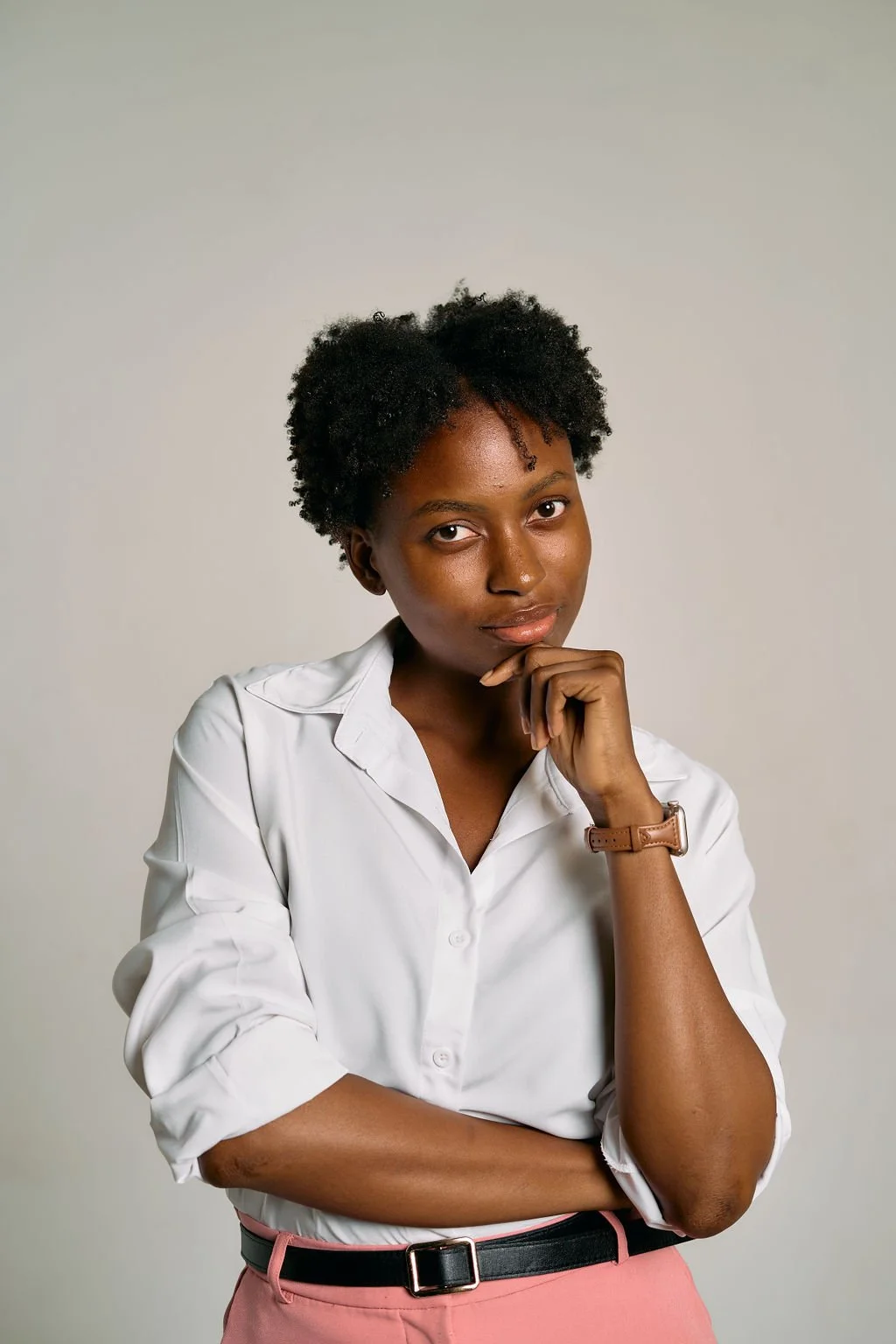 A woman with natural curly hair wearing a white button-up shirt and pink pants, resting her chin on her hand, looking at the camera.