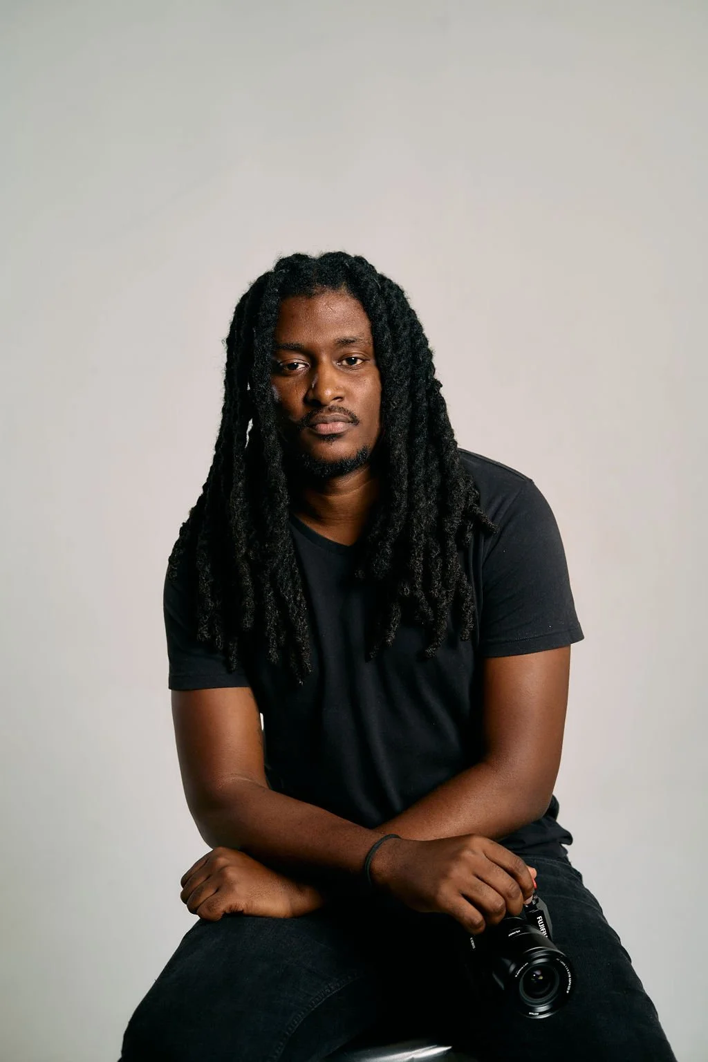Portrait of a man with long dreadlocks wearing a black t-shirt, holding a camera, sitting against a plain white background.