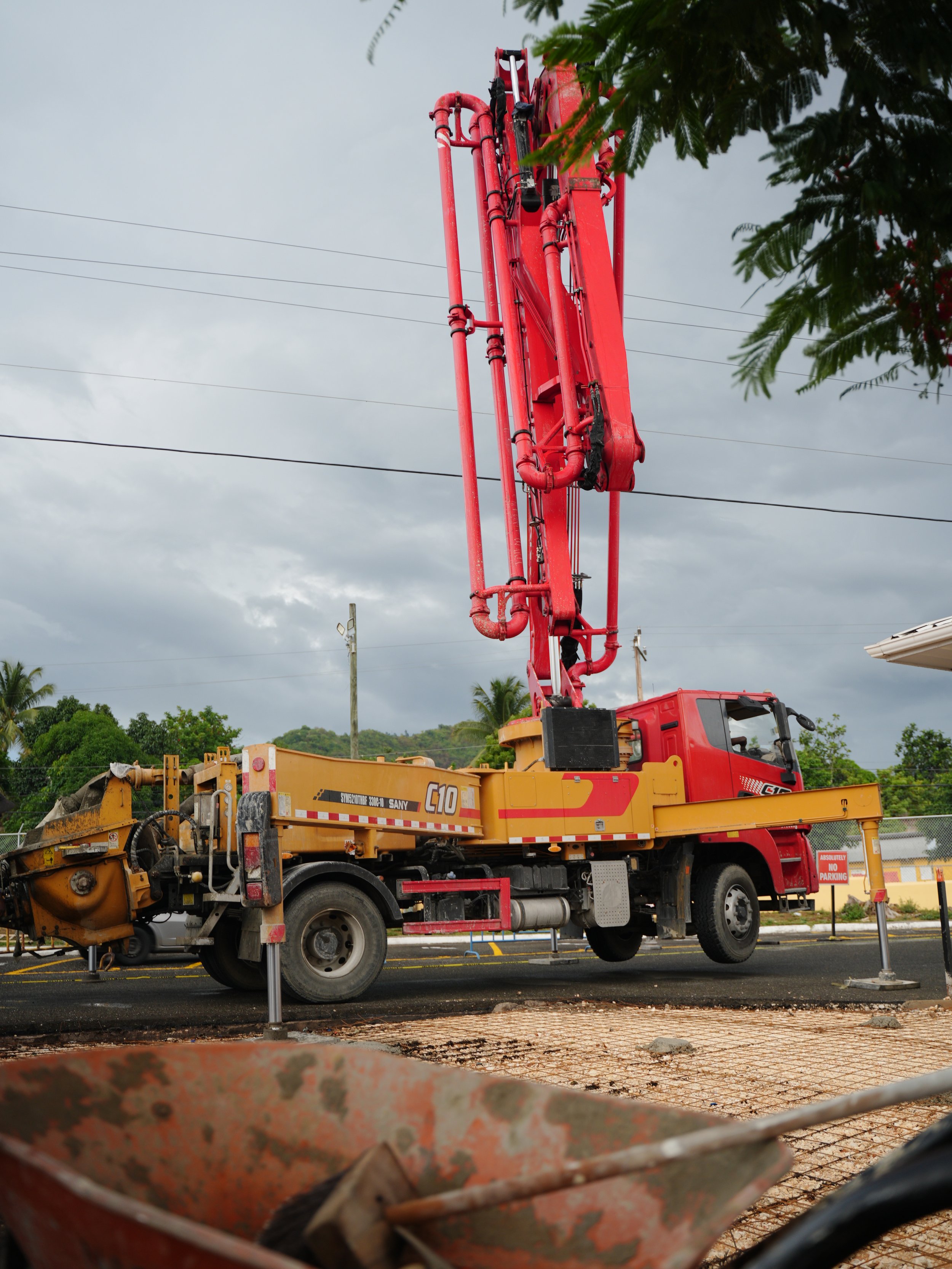 A red and yellow roadside construction crane with a long arm extended upwards, on a cloudy day, with trees and electrical wires in the background.