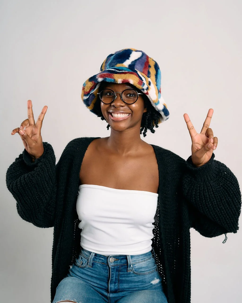 Young woman smiling and making peace signs with both hands, wearing a colorful hat, glasses, a white tube top, a black cardigan, and blue jeans against a plain background.