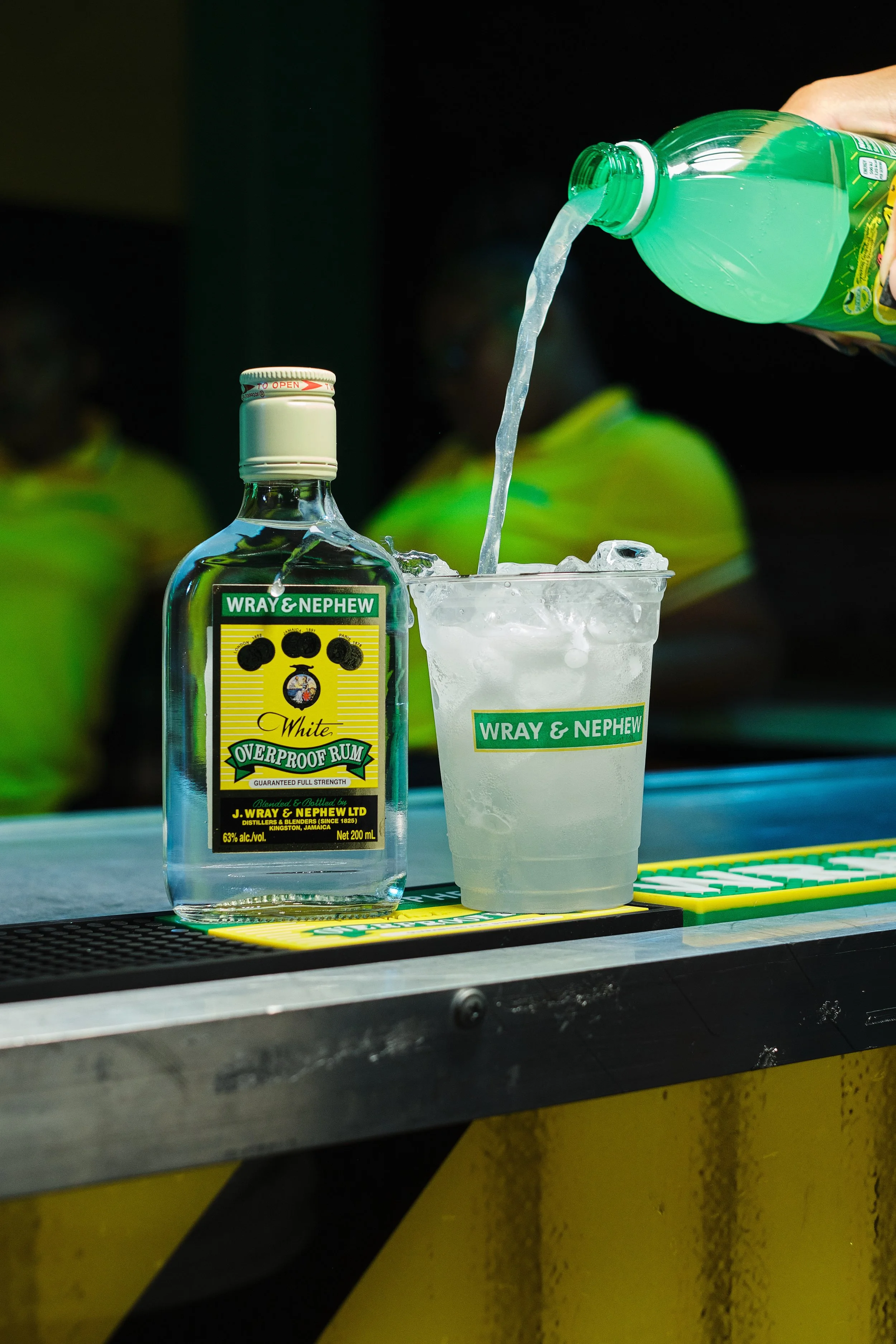 A bottle of Wray & Nephew white overproof rum and a plastic cup filled with ice, with a hand pouring a green soda into the cup.