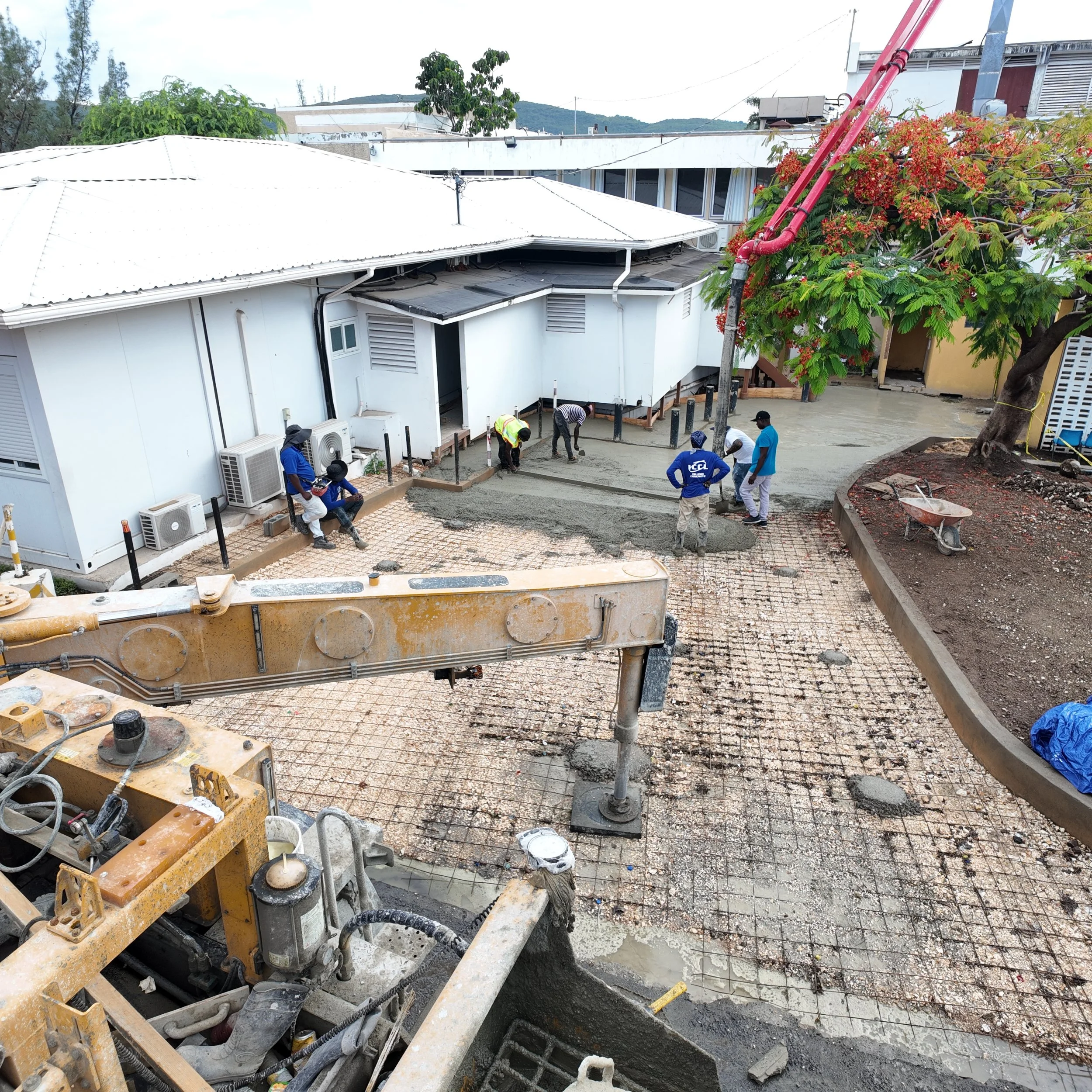 Construction workers laying concrete in a courtyard area with a white building and a tree with red flowers nearby.